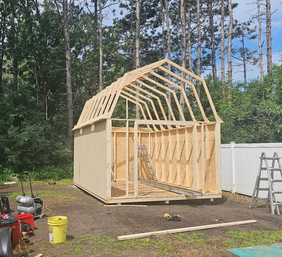 A wooden barn-style shed under construction in a backyard with visible framing and unfinished siding. A ladder and tools are nearby.