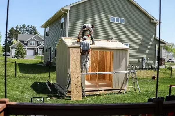 Two people constructing a shed in a backyard, with green siding and a house in the background on a sunny day.