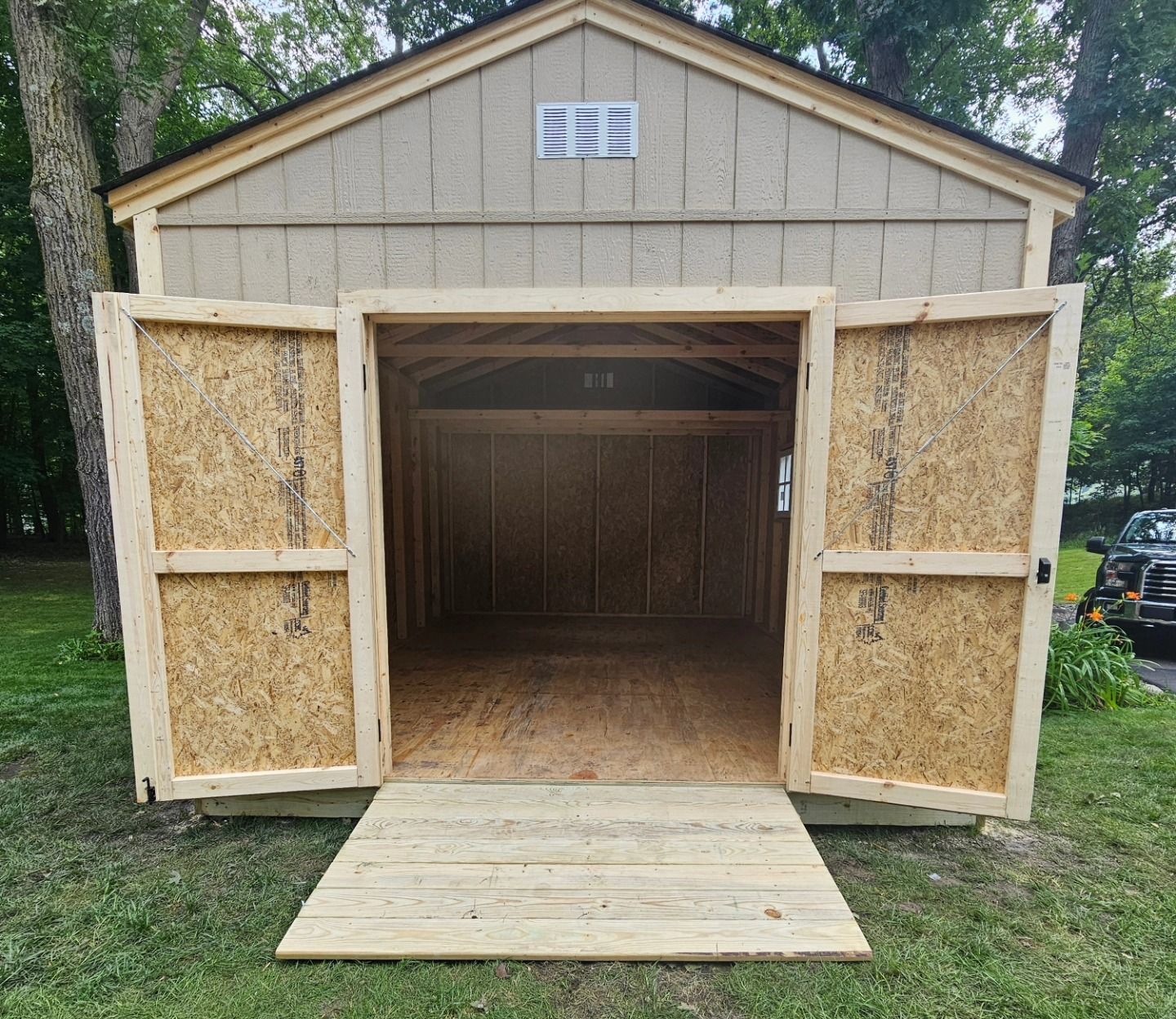 A wooden shed with open double doors and a ramp, set on grass. The shed is empty inside.