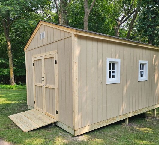 Tan shed with a ramp, two windows, and double doors, set on a grassy lawn.
