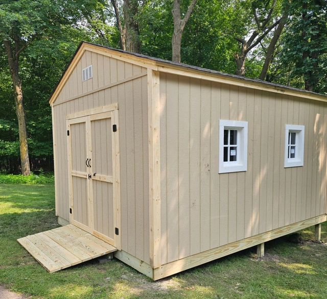 Tan shed with a ramp, two windows, and double doors, set on a grassy lawn.