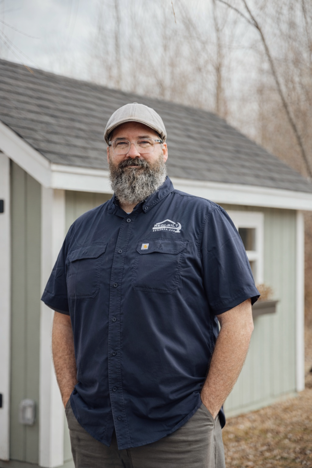 A bearded man in a blue shirt and cap stands in front of a small shed, hands in pockets.