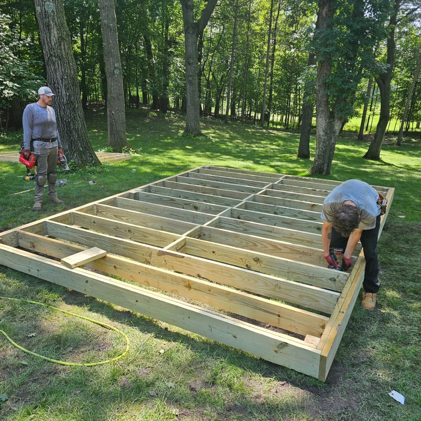 Two men building a wooden deck in a grassy, wooded area. One is standing, the other is working with a drill.