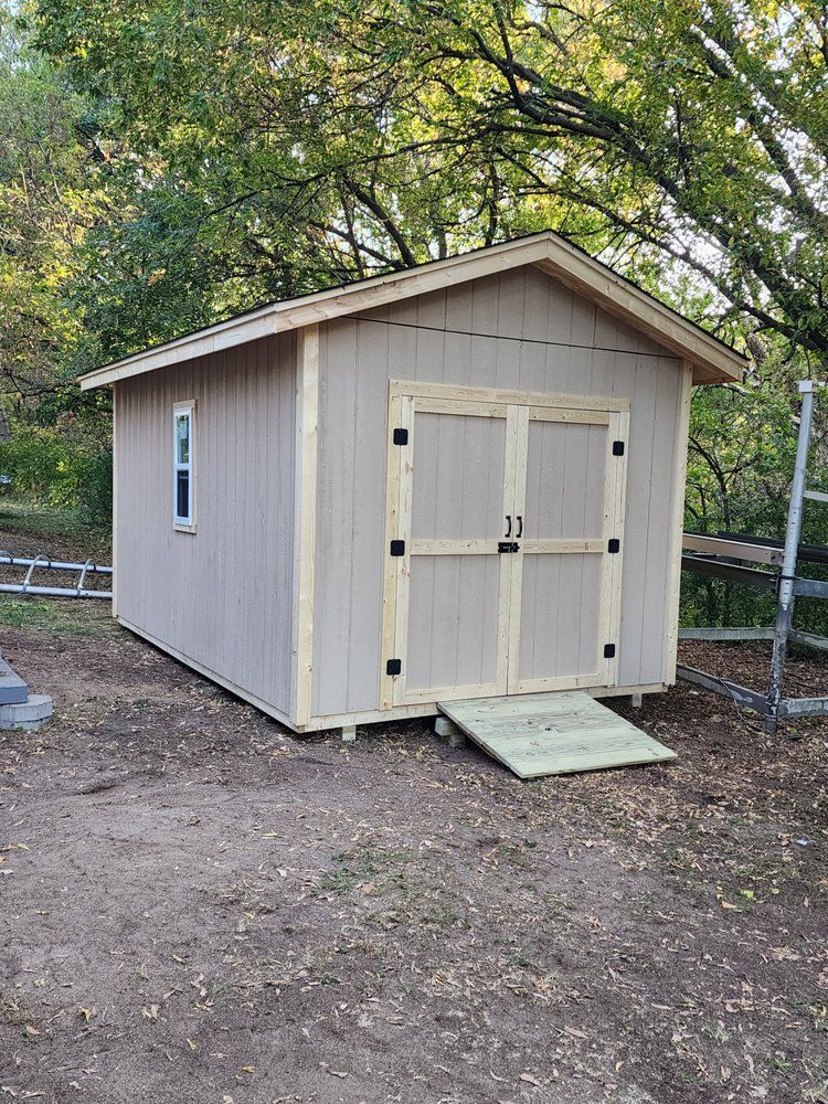 Tan shed with double doors and ramp, beige trim, small window, and green foliage.