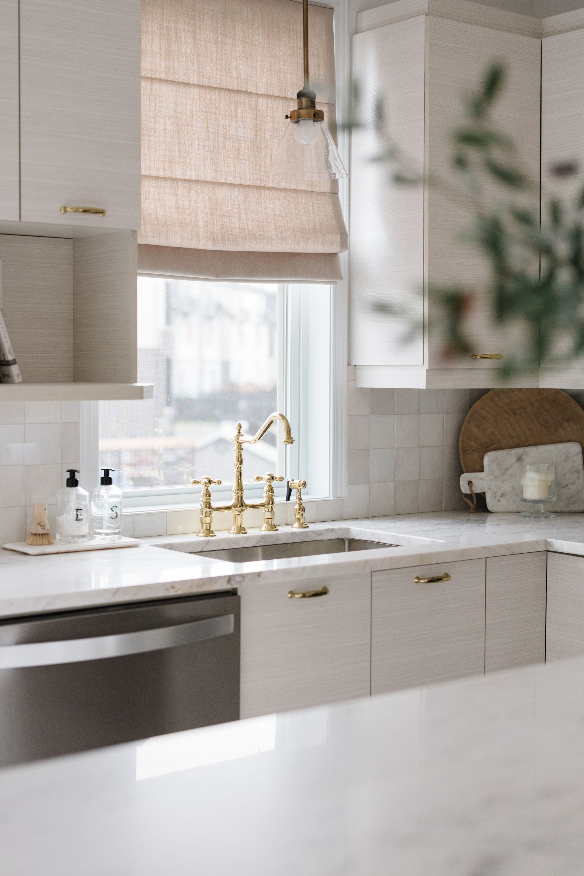 White kitchen with gold faucet, tan window shade, and stainless steel dishwasher.
