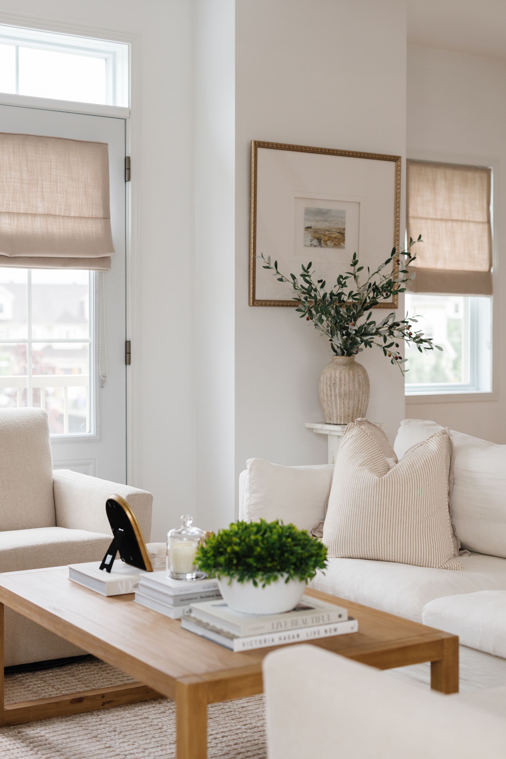 Living room with neutral tones; white walls, tan shades, wooden coffee table, sofa with pillows, and framed art.
