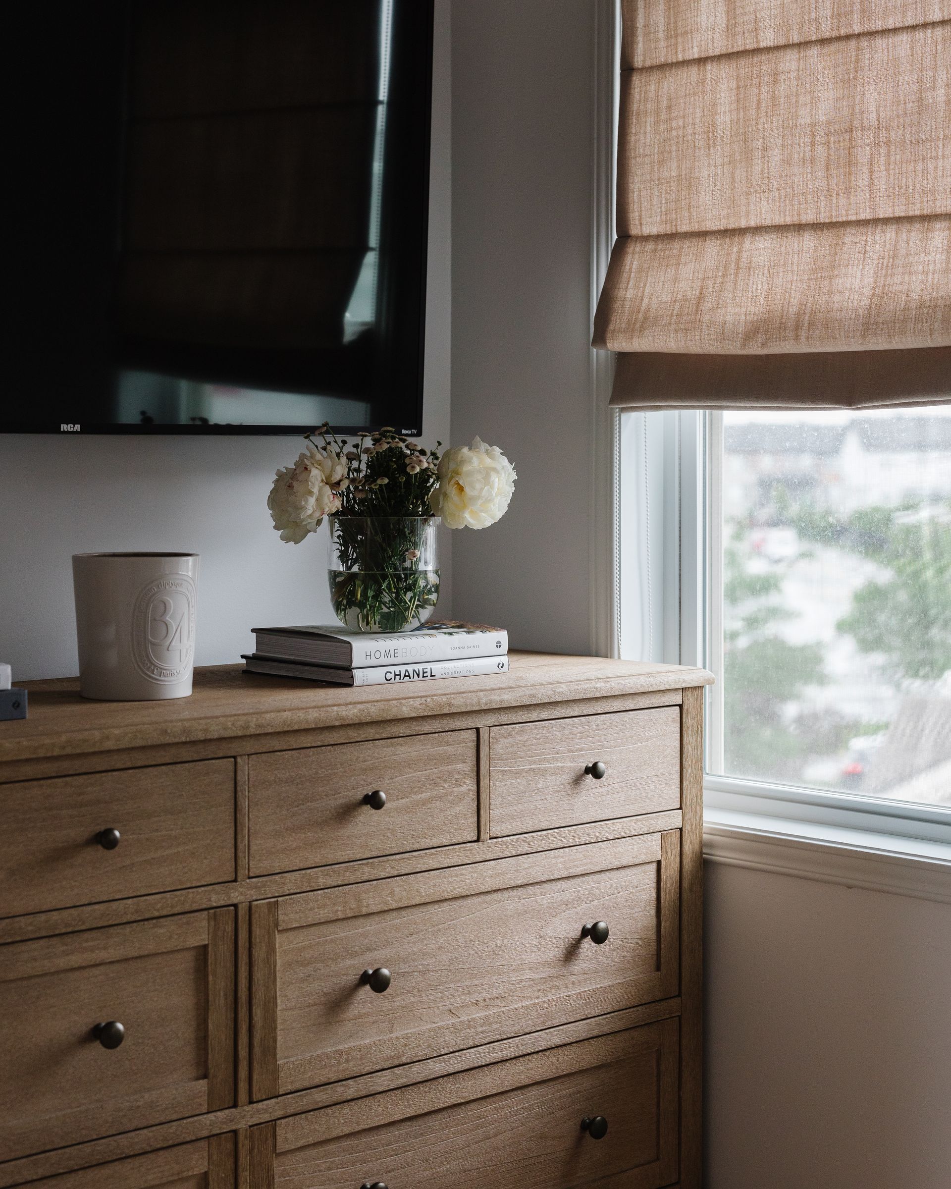 Wooden dresser with TV, flowers, and window with a beige Roman shade.