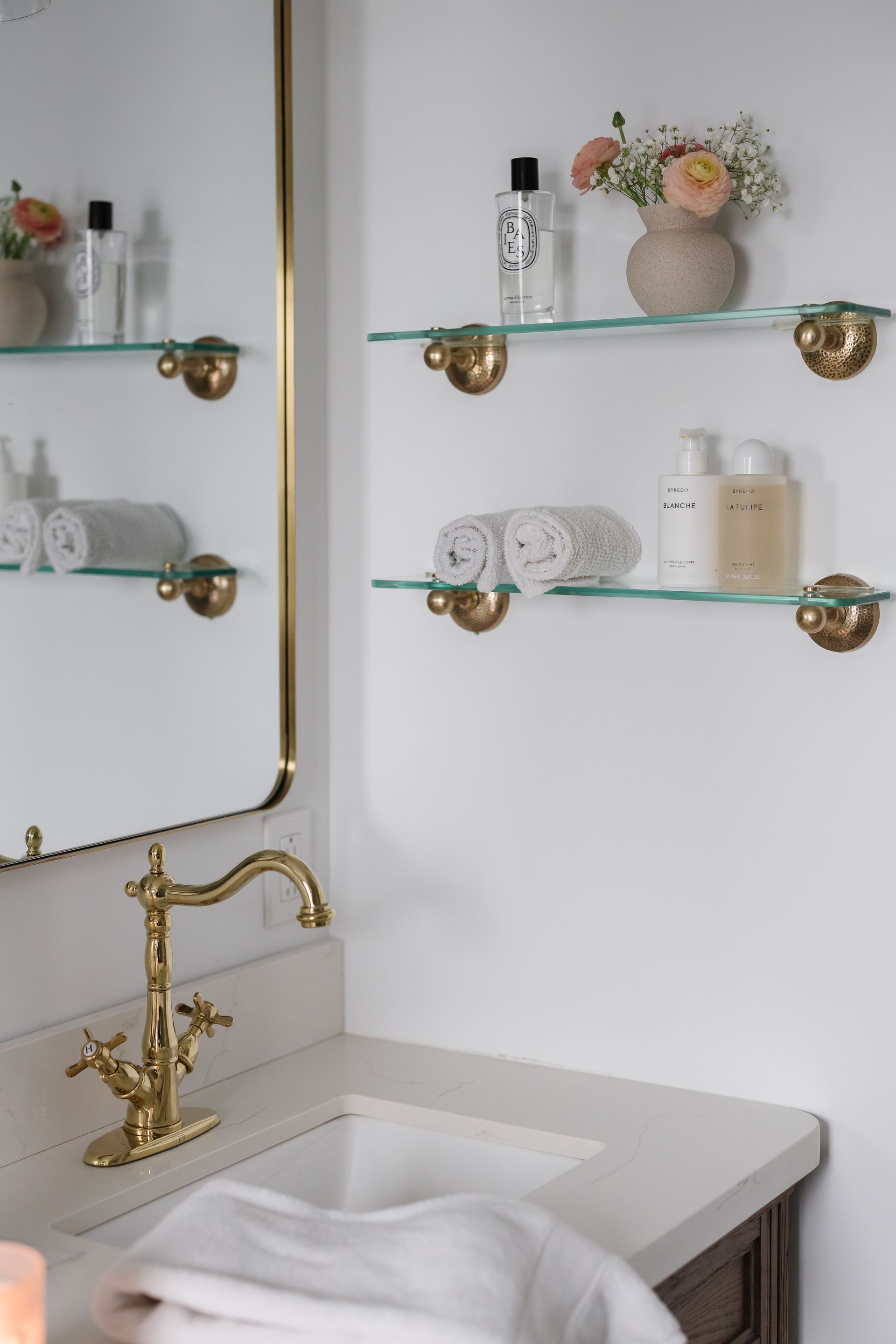 Bathroom with glass shelves and gold fixtures, holding toiletries and a vase of flowers.