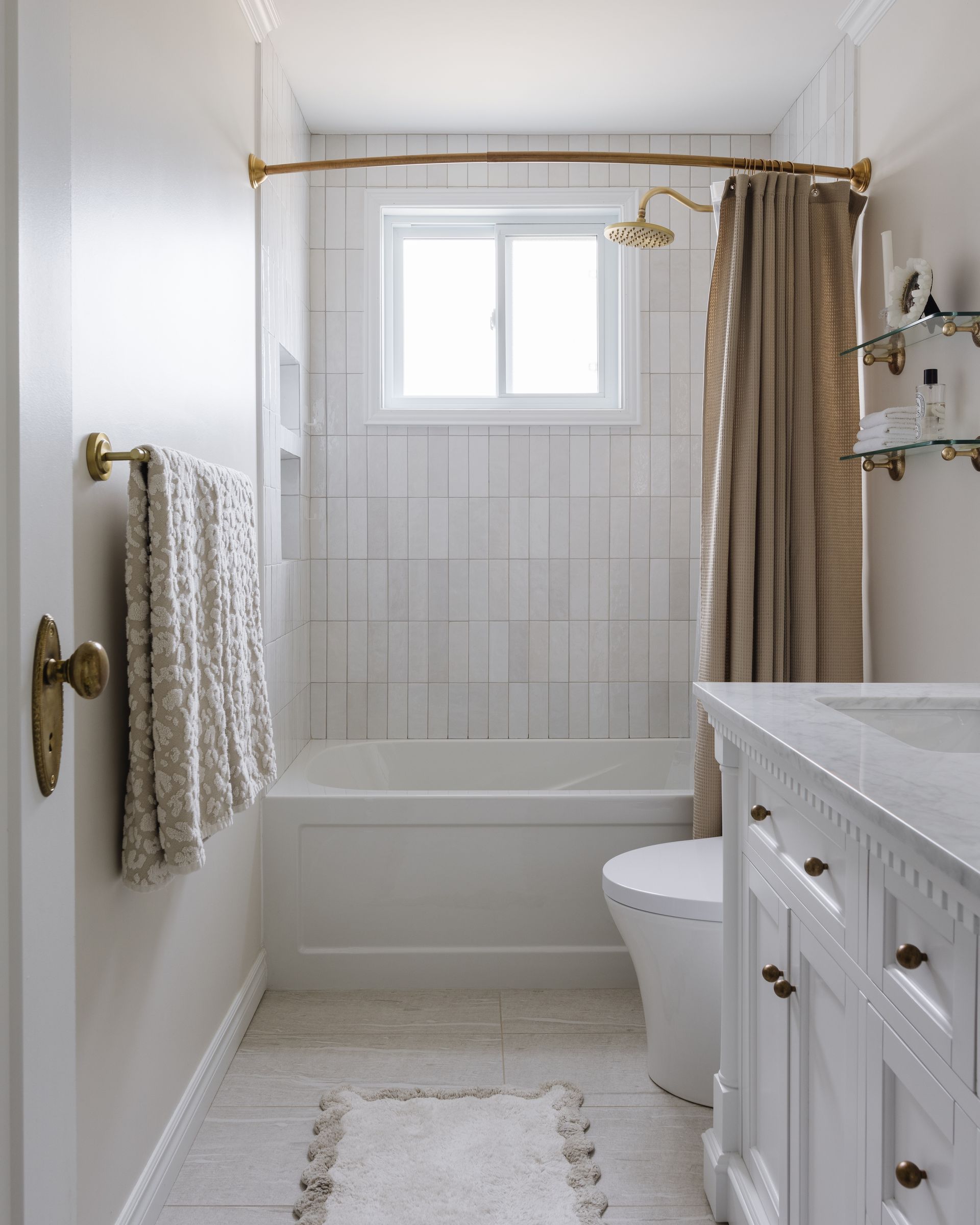 Narrow white bathroom with a bathtub, toilet, and vanity; gold fixtures and a beige curtain.
