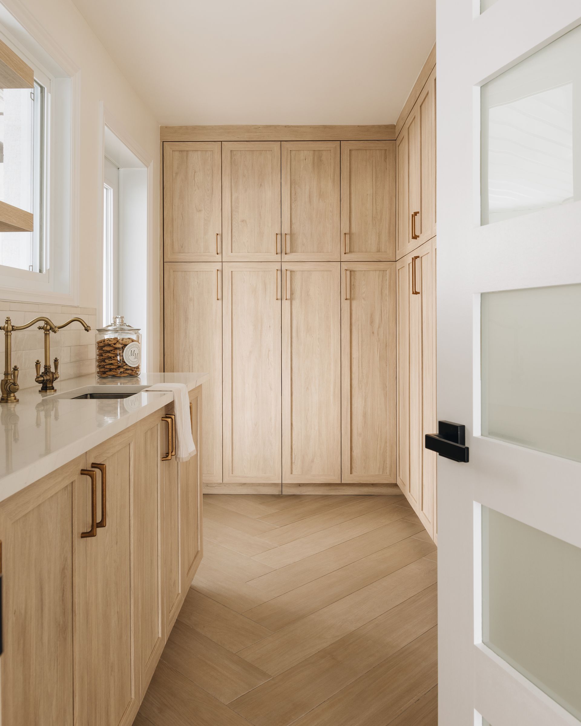 Laundry room with light wood cabinets, white countertops, brass fixtures, and a herringbone floor.