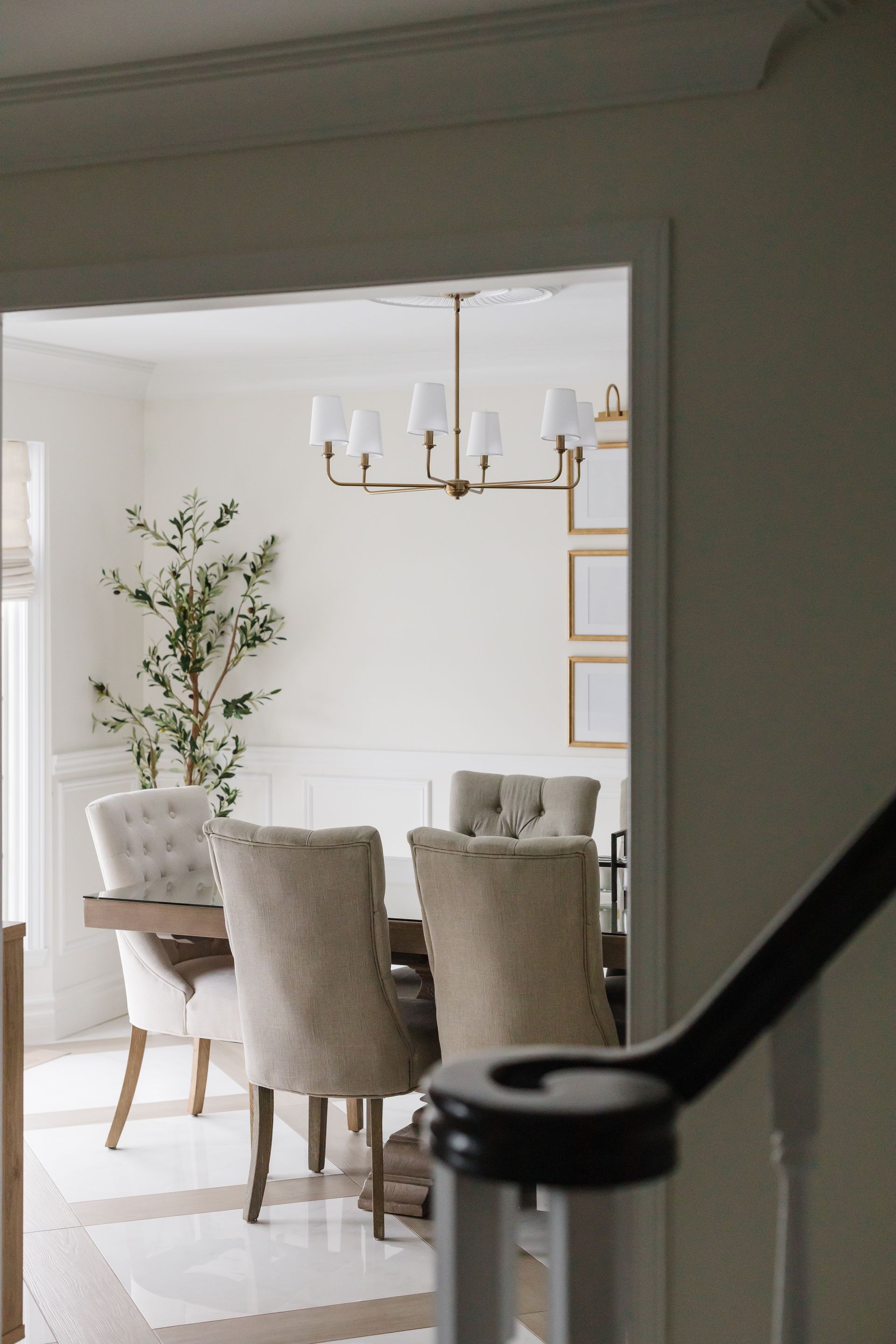 Dining room interior, seen through doorway. Beige chairs surround a wooden table. Chandelier hangs above.