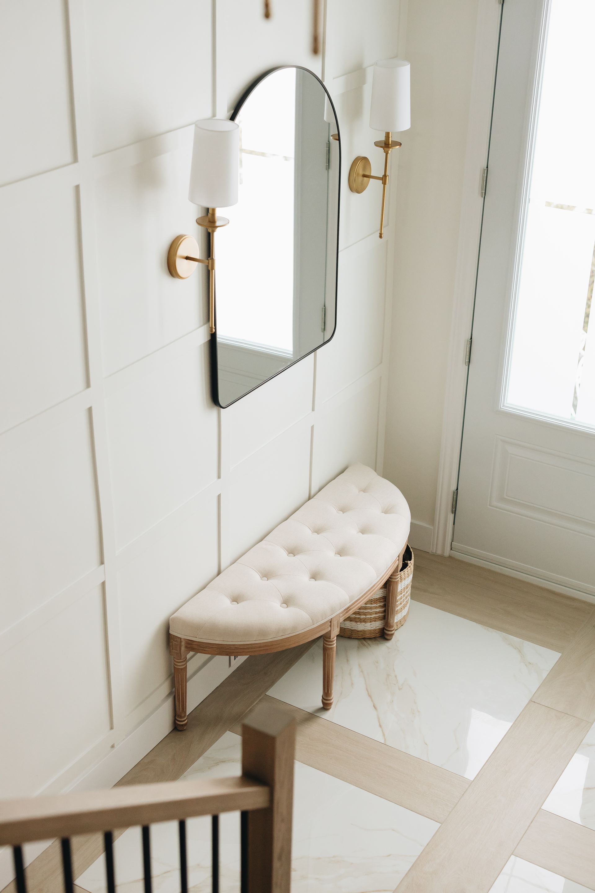 Entryway with mirror, bench, sconces, and white paneled wall.