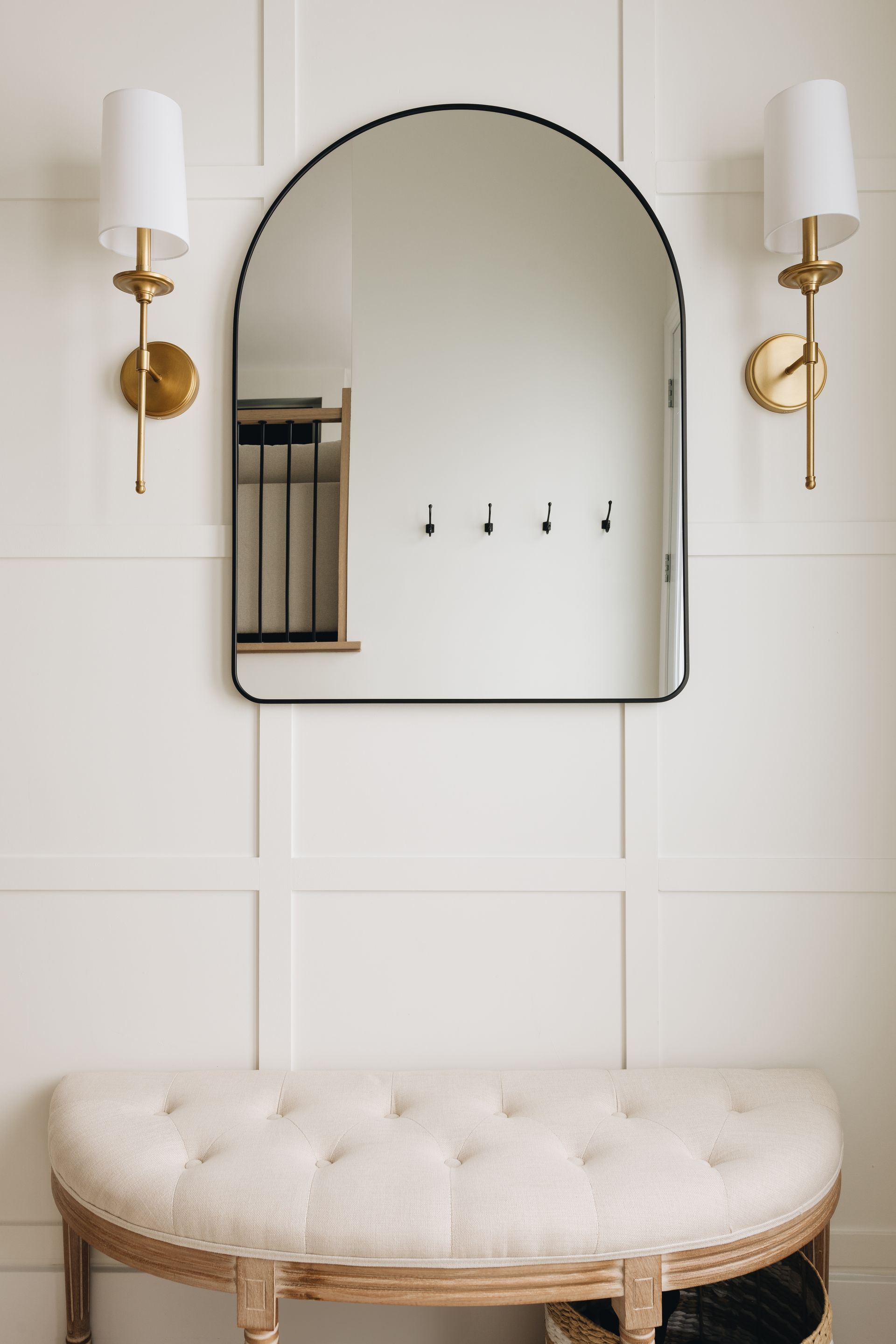 Entryway with arched mirror, sconces, and tufted bench against paneled wall.