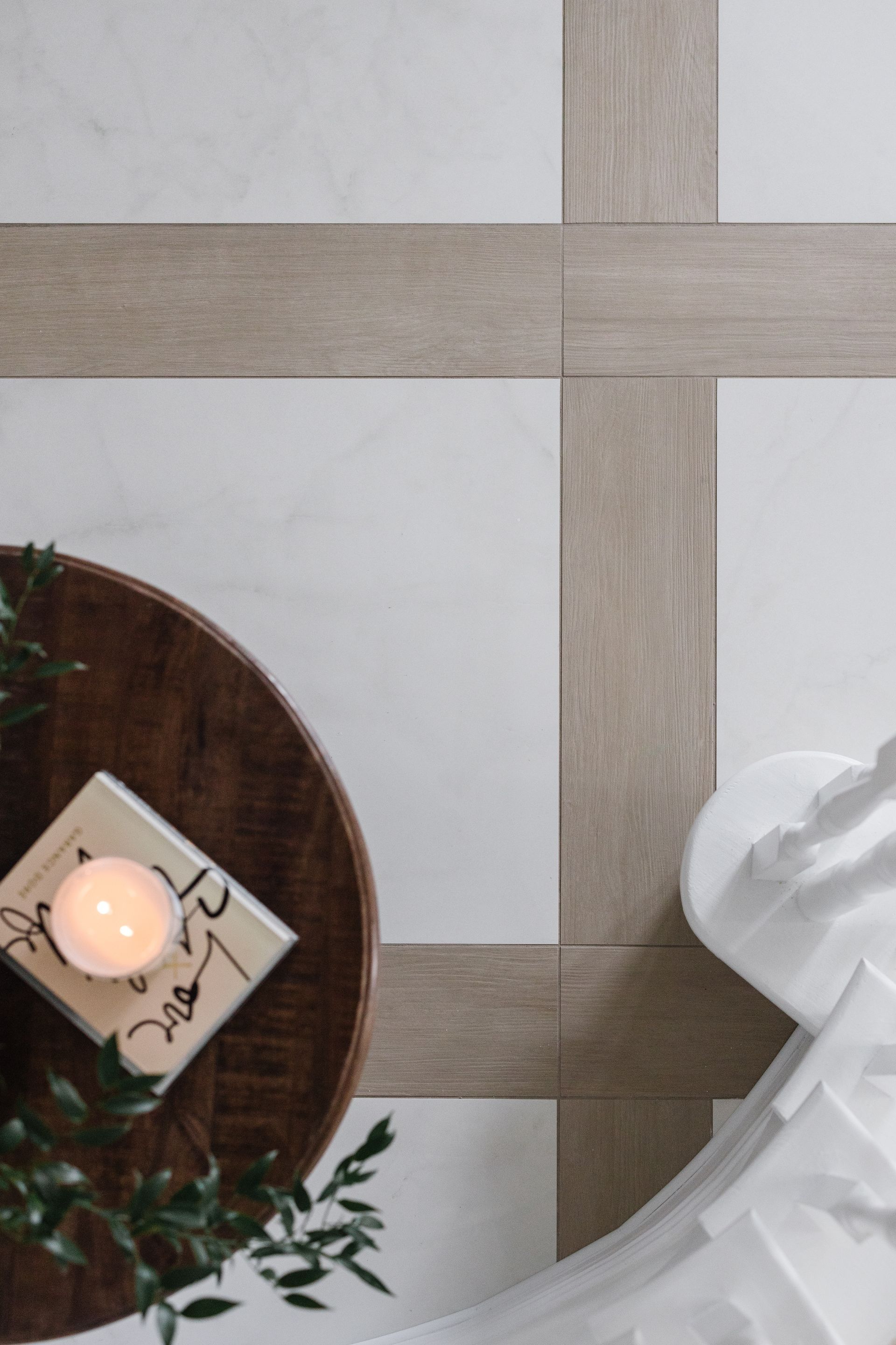 Overhead view of a wooden tray with a candle and book on patterned white and wood floor tiles.