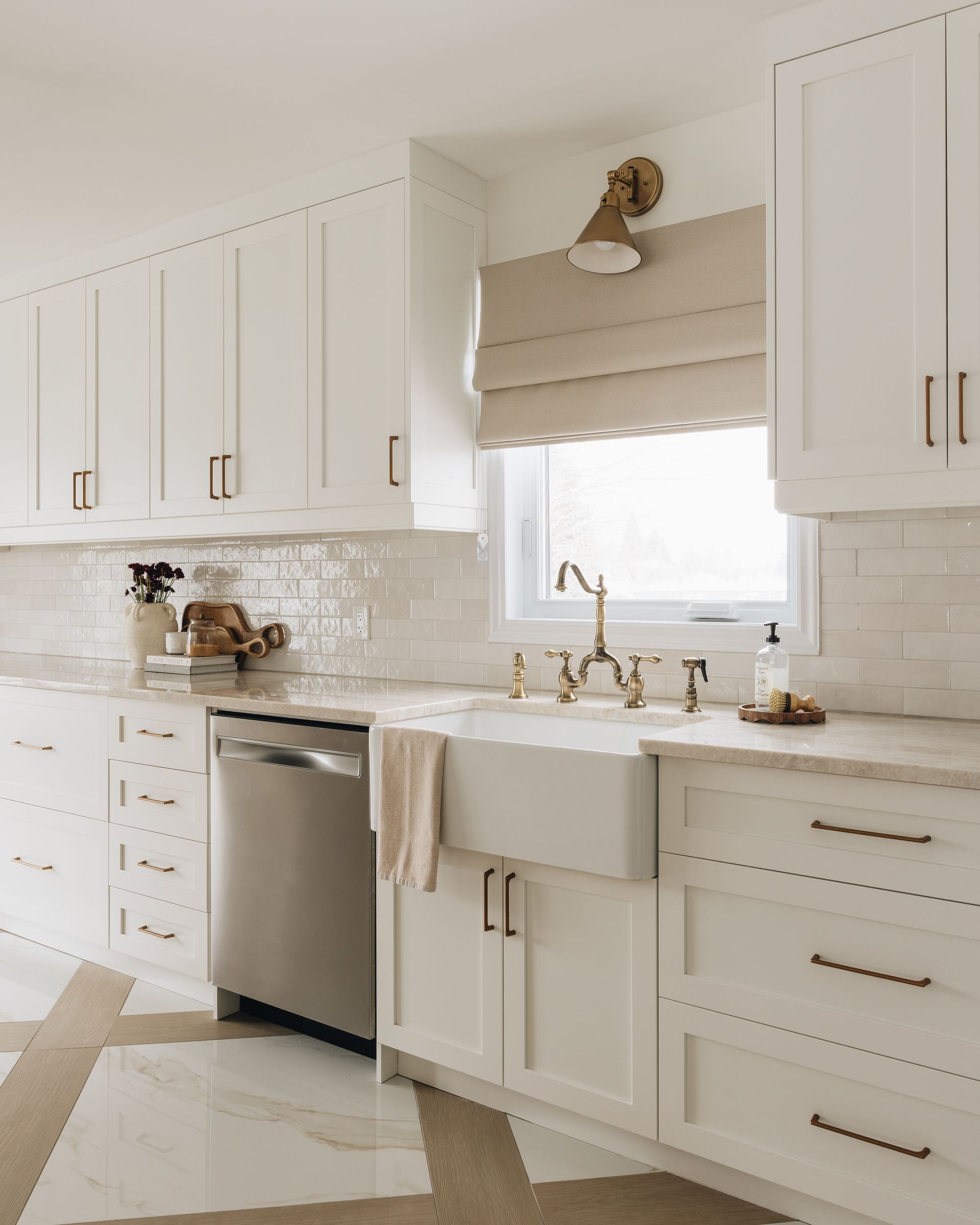 White kitchen with cabinetry, stainless steel dishwasher, farmhouse sink, brass faucet, and window.