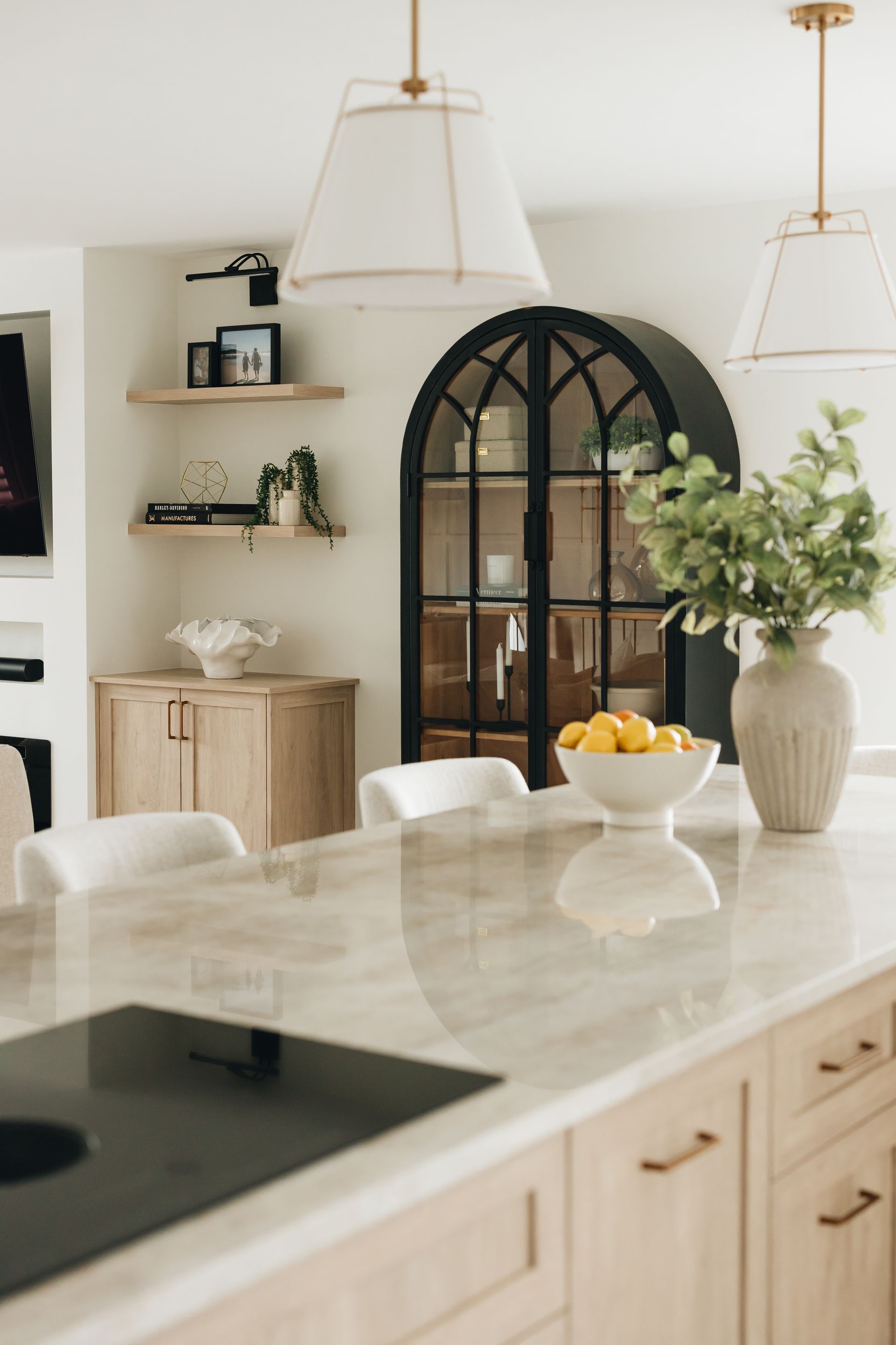 Elegant kitchen with light wood cabinets, black arched cabinet, floating shelves, and island with a cooktop.