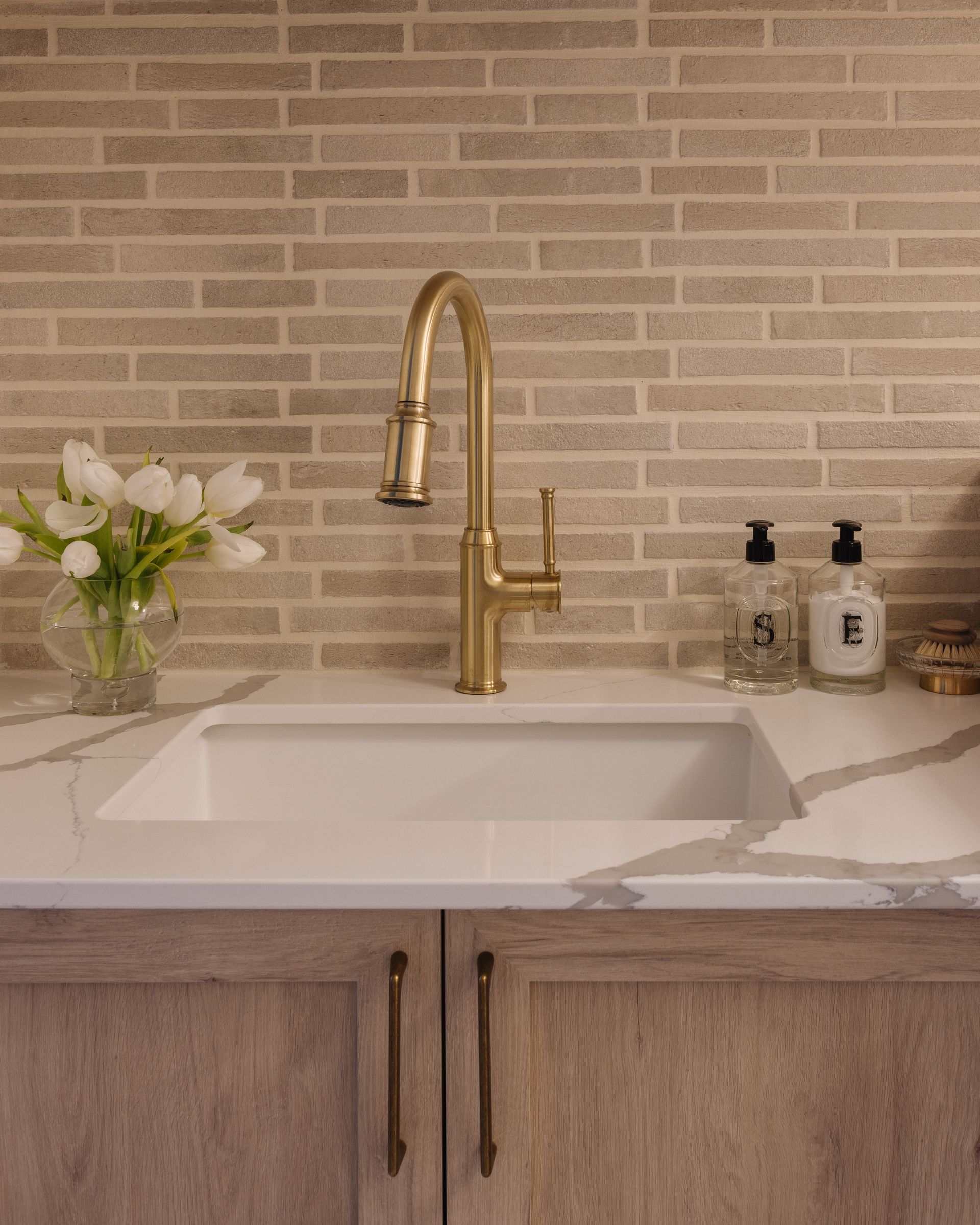 Kitchen sink with gold faucet, white countertop, and beige tile backsplash. Cabinet doors below.