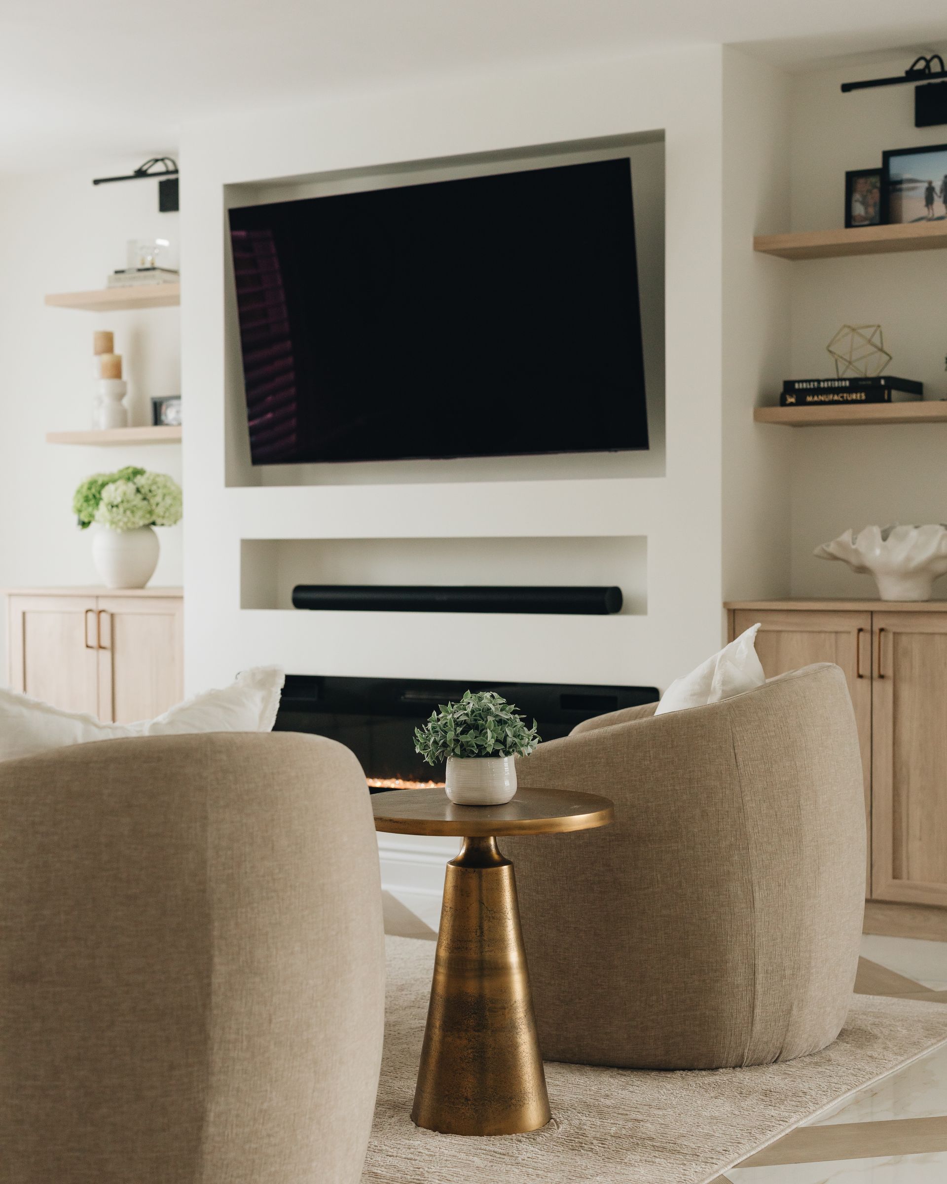 Living room with TV above fireplace, beige chairs, gold side table, and shelves with décor.
