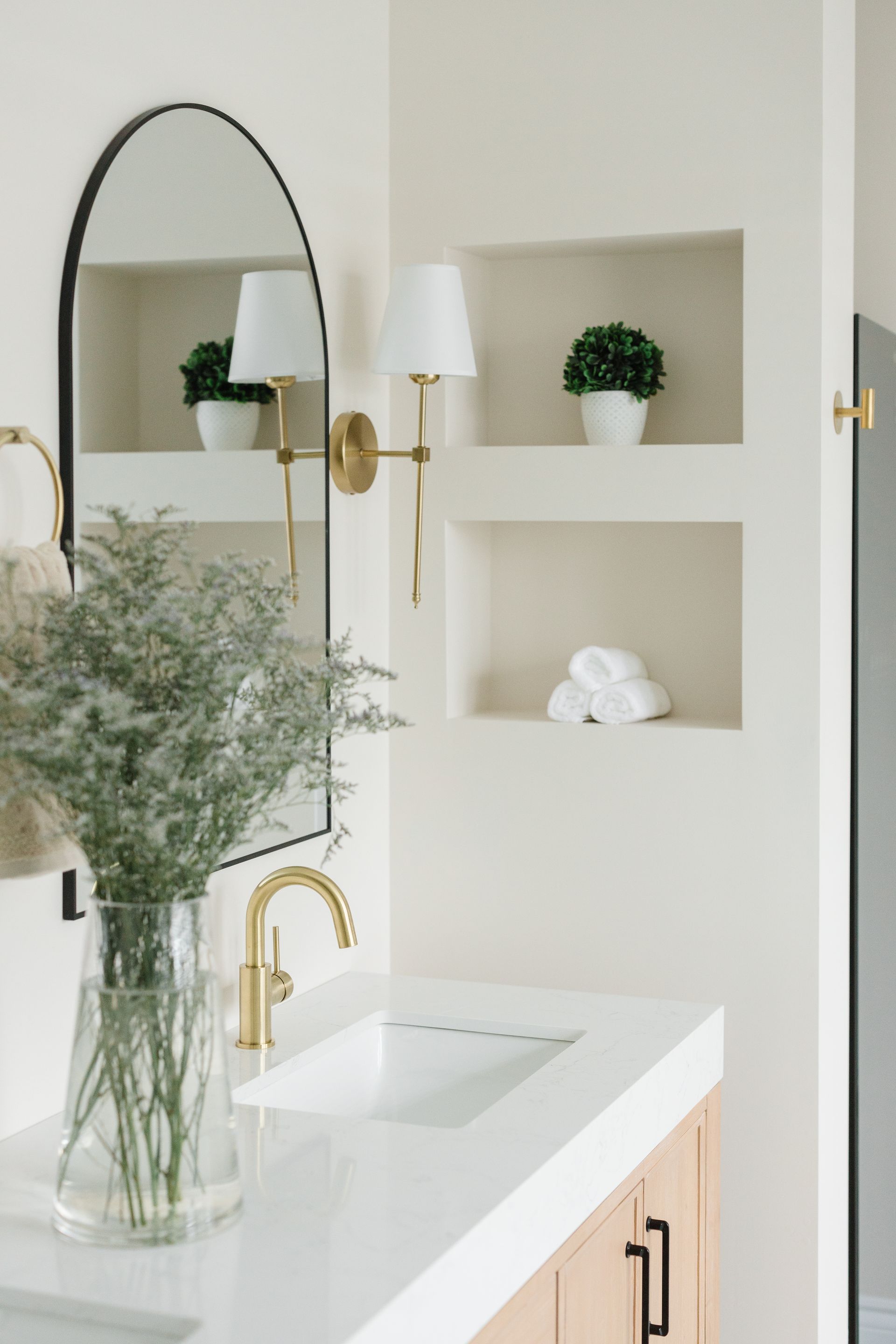 Bathroom with a white countertop, gold fixtures, and an arched mirror. Built-in shelving displays plants and towels.