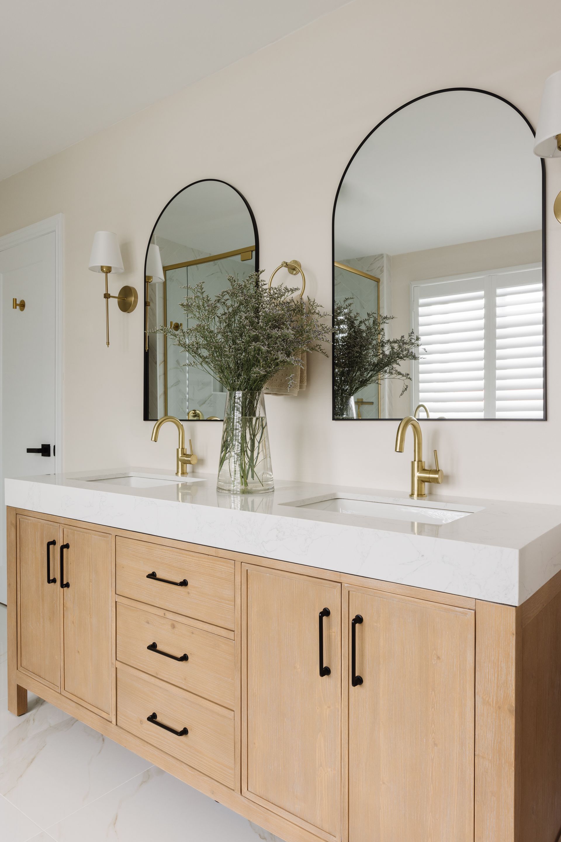 Bathroom with light wood vanity, marble countertop, arched mirrors, and gold fixtures.