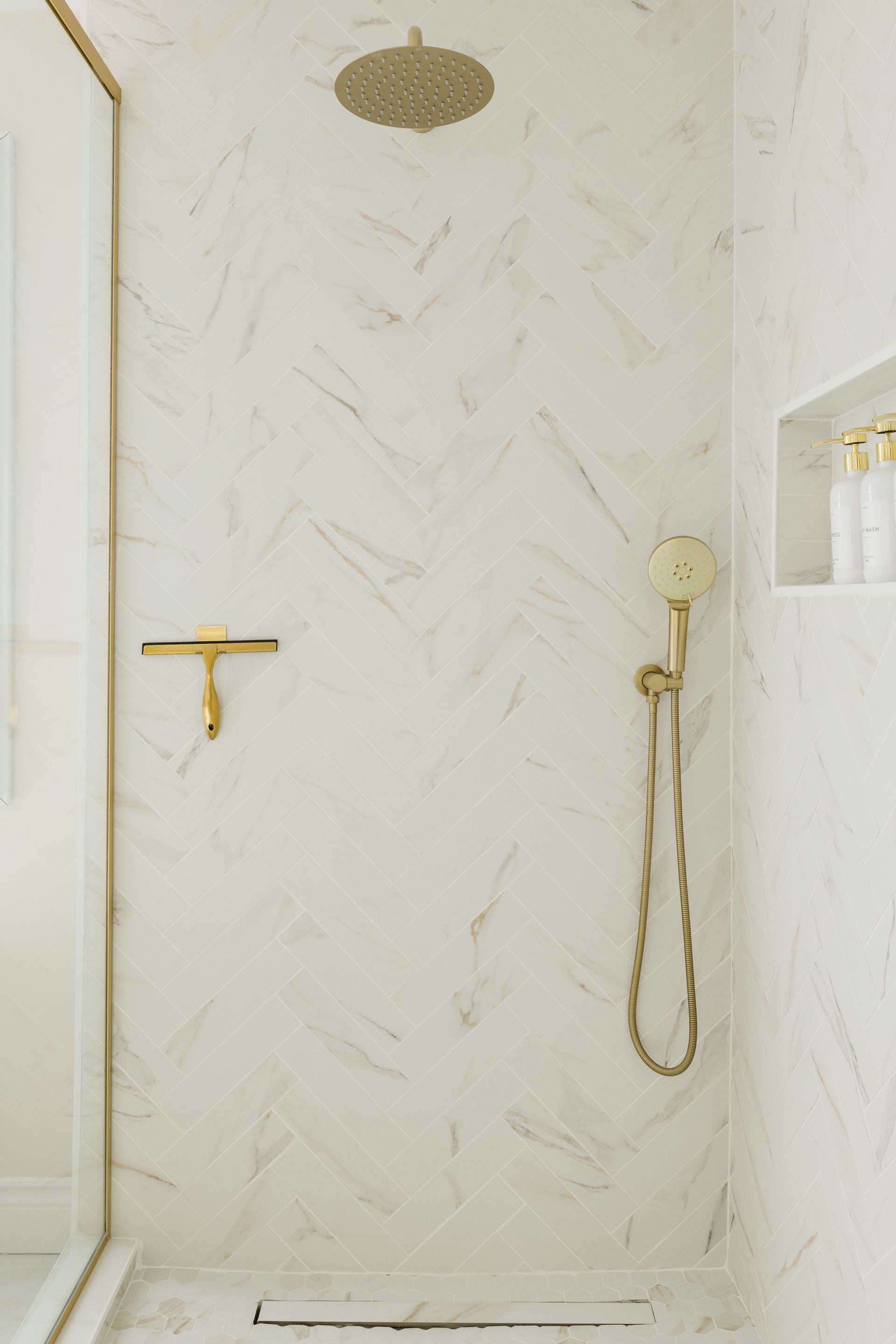 A modern shower with white marble walls, gold fixtures, and a glass door.