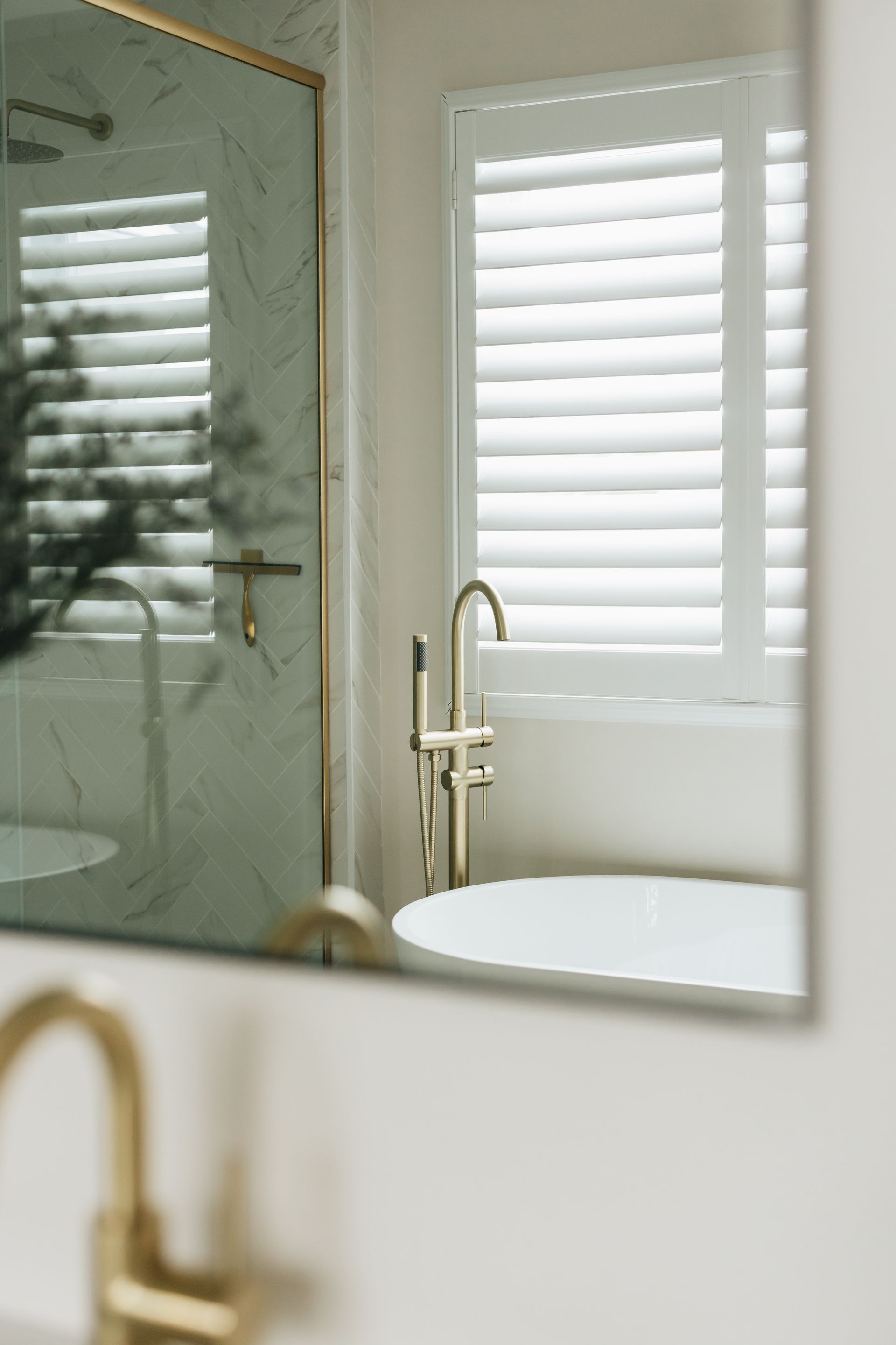 Mirror reflection of a bathroom with gold fixtures, a white bathtub, and shutters on the window.