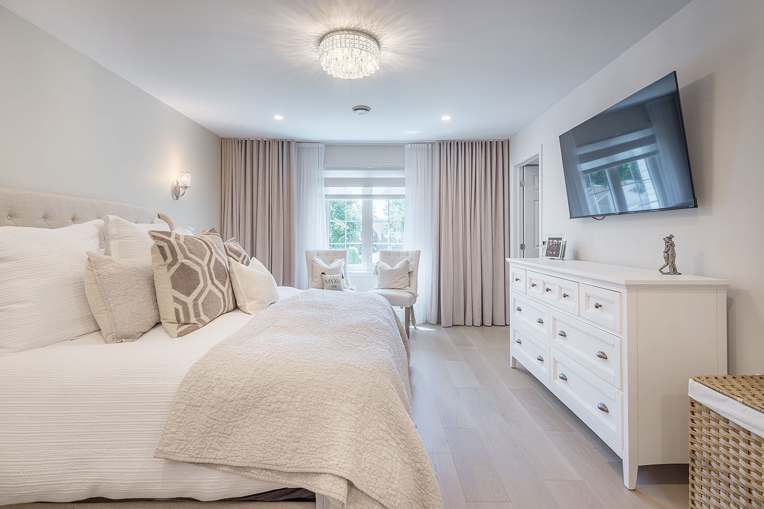 Bedroom with a white dresser, bed, and mounted TV. Neutral colors, light wooden floor, and curtains.