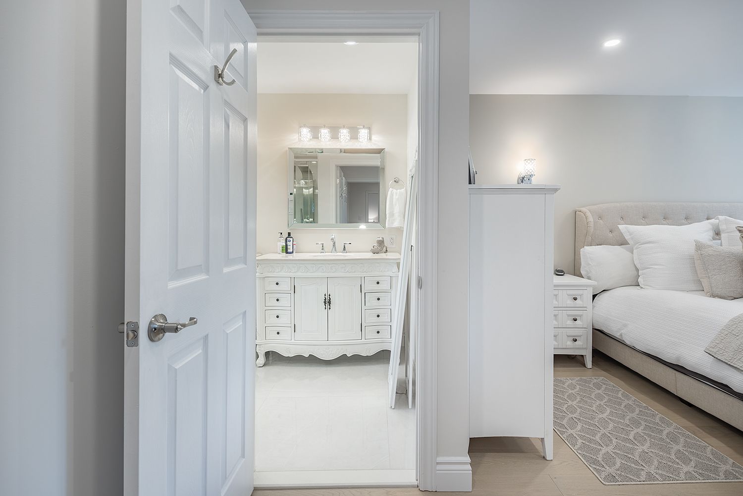 White bathroom visible through an open door from a bedroom with a bed and nightstand.