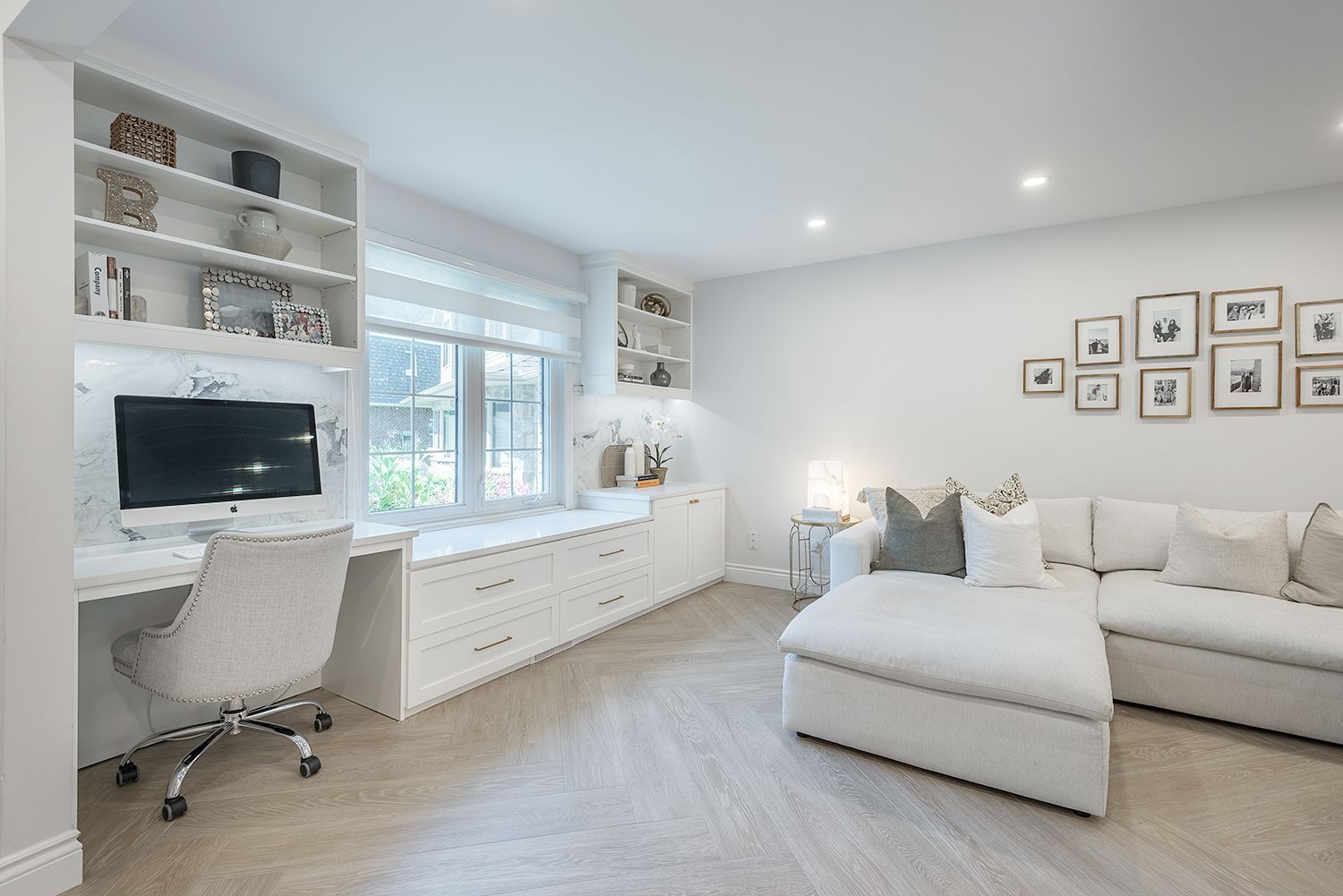 White home office with built-in desk, shelving, and a sofa. Light flooring and decor.