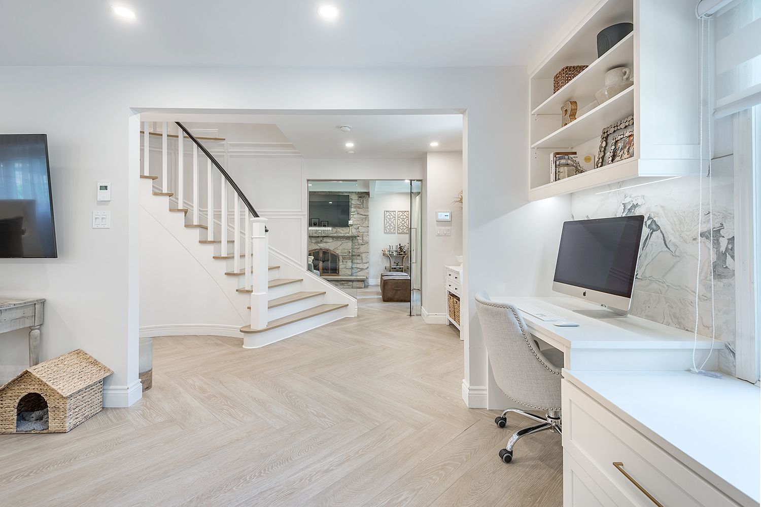 White home office with built-in desk, shelving, stairs, and doorway leading to a living room with a fireplace.