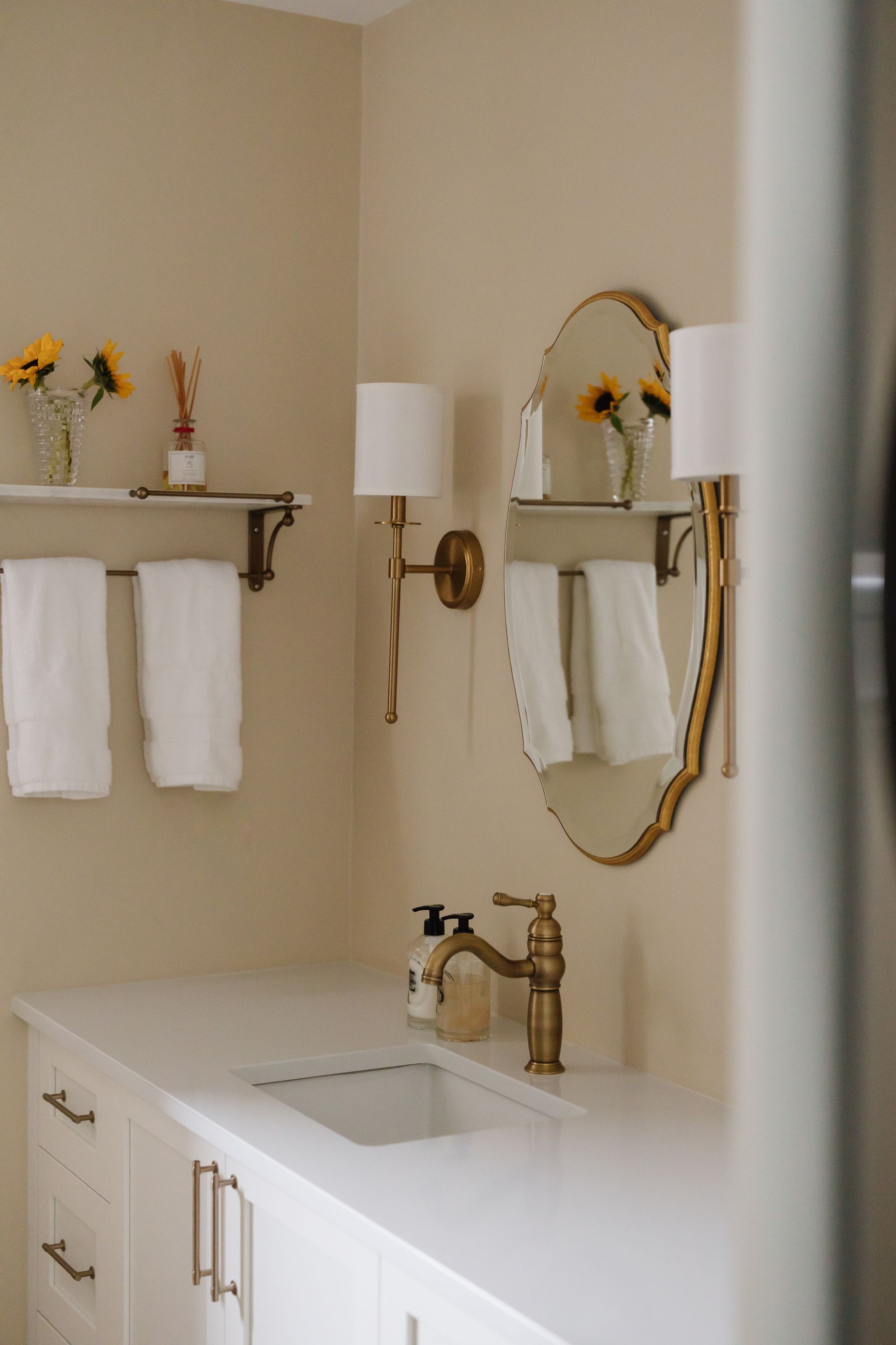 Bathroom with white countertop, brass fixtures, and gold-framed mirror; sunflowers in vases.