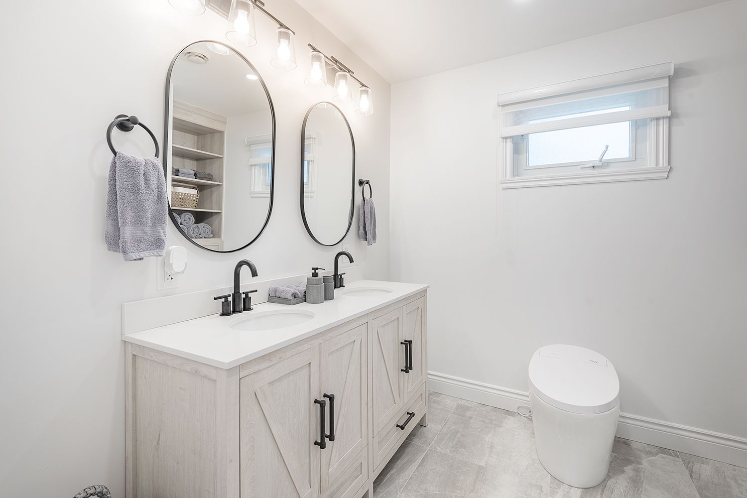 Bathroom with double vanity, oval mirrors, and white toilet. Gray and white color scheme.