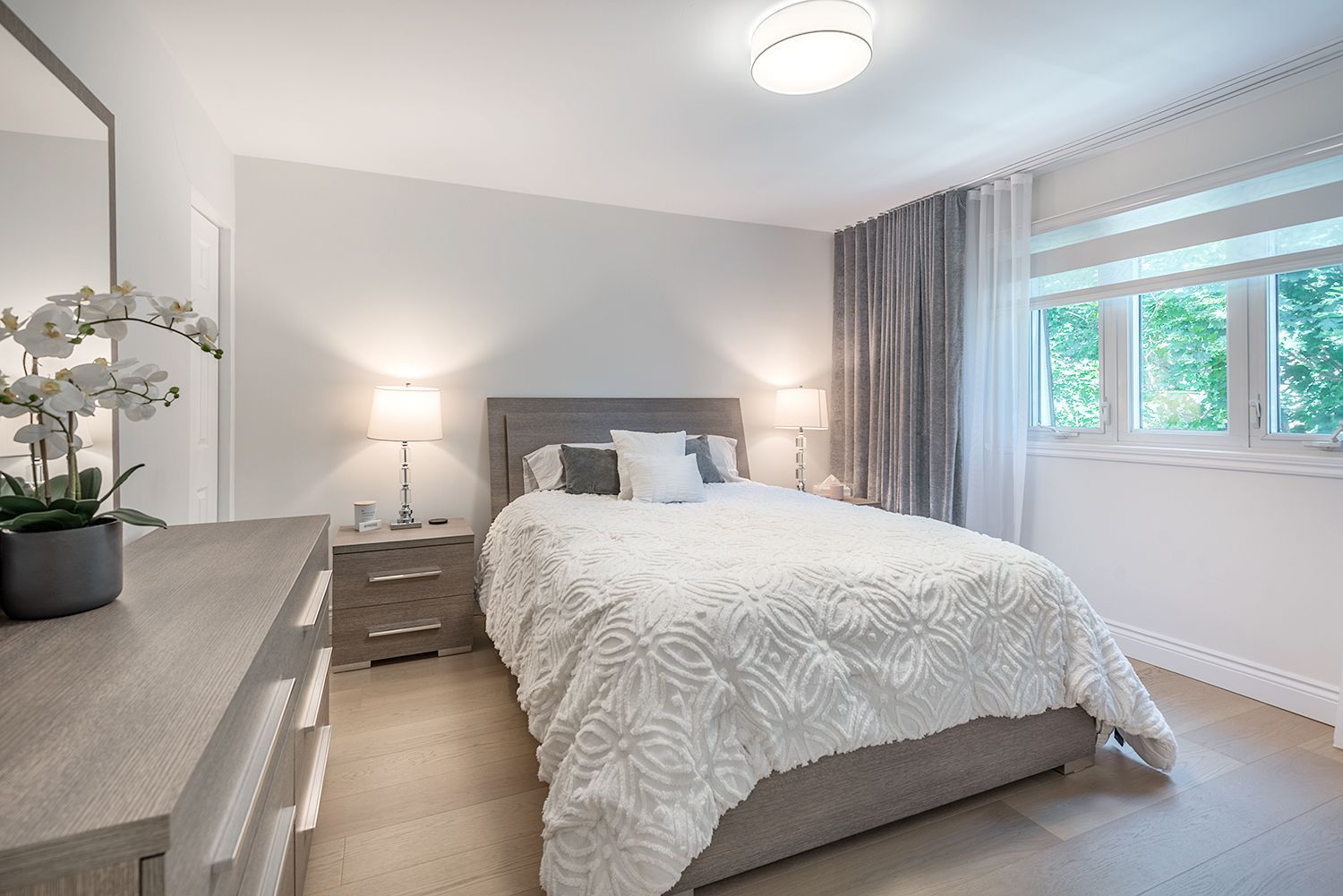 Bedroom with a gray bed, white bedding, nightstands, a dresser, and a window with sheer curtains.