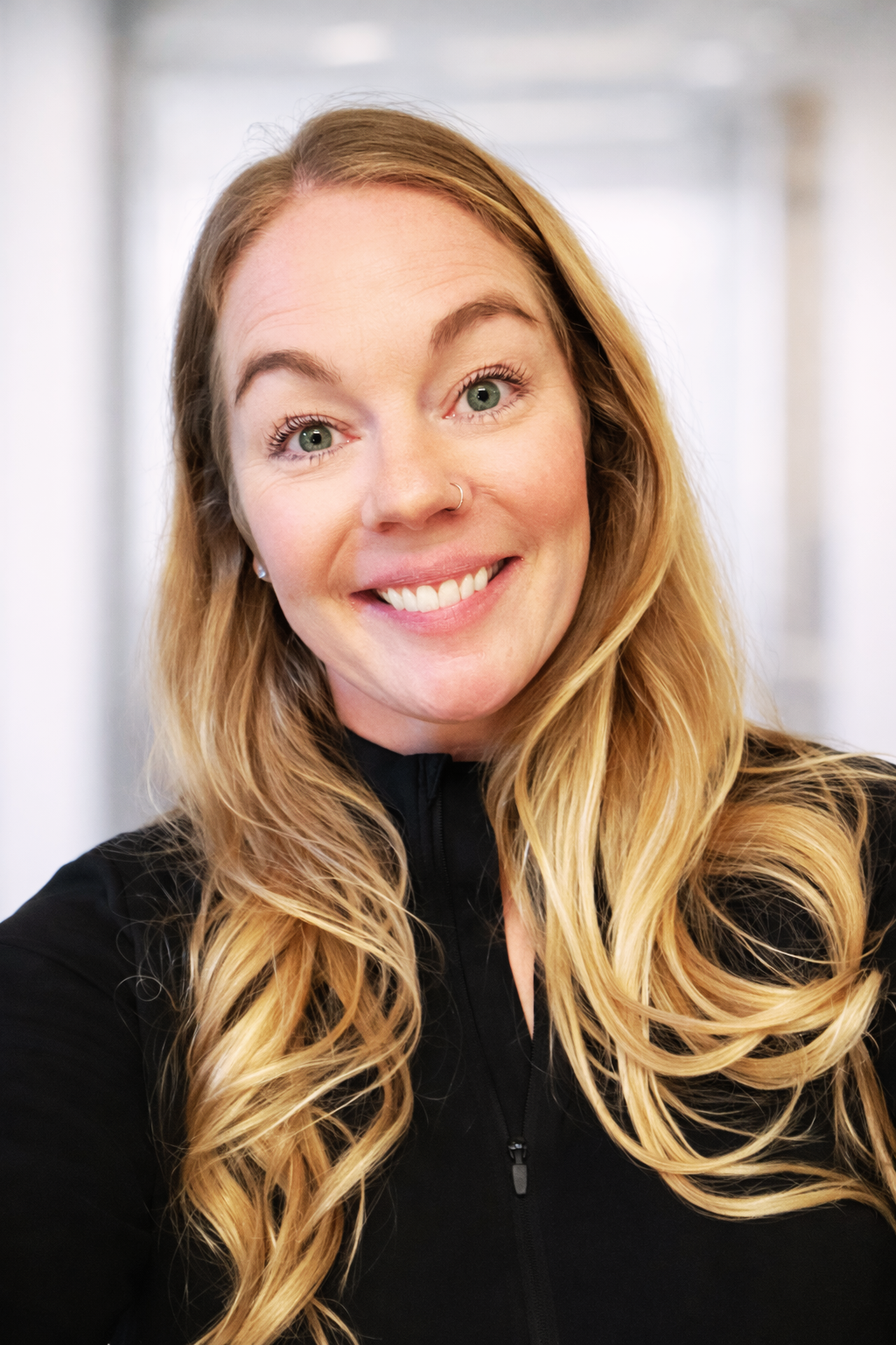 Smiling woman with long blonde hair in a black top, standing indoors against a blurred light background