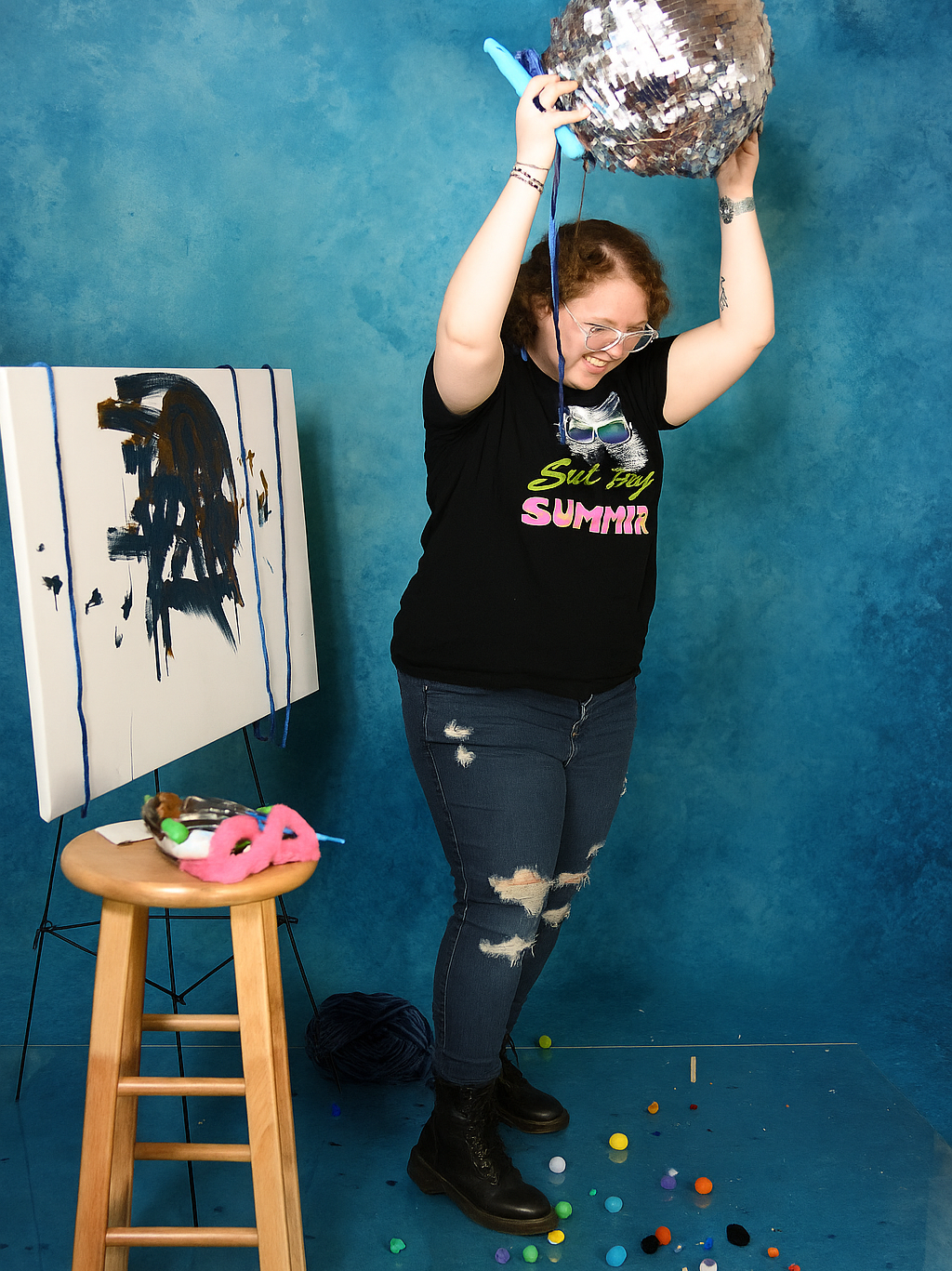 Person smashing a disco ball with a stick, art easel and stool present, colorful items scattered.