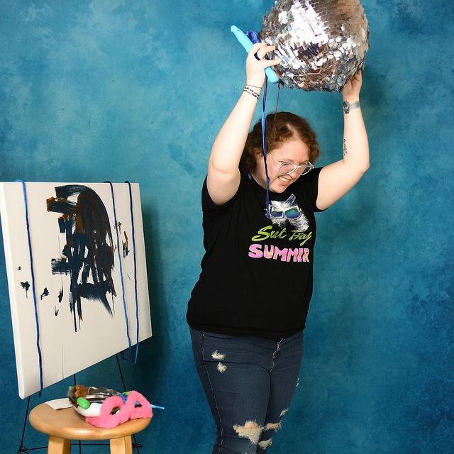 Person painting a canvas with an art easel and stool present, colorful items scattered.