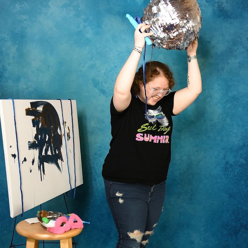 Person painting a canvas with an art easel and stool present, colorful items scattered.