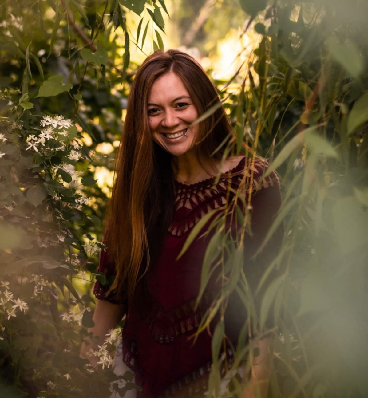 Woman with long hair smiles, framed by leafy foliage, wearing a burgundy embroidered top in a forest.