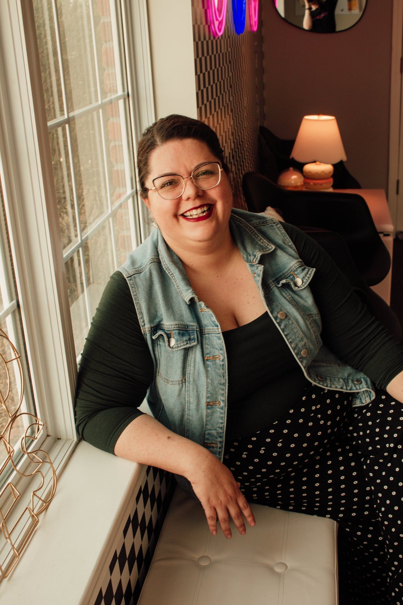 Smiling woman sitting by a window, wearing glasses, a green vest, and a black polka-dot skirt.