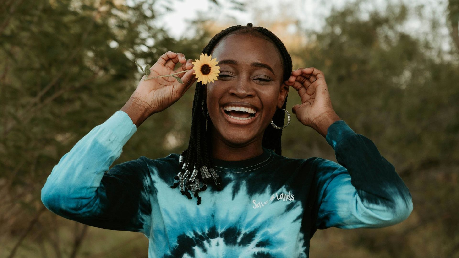 A woman in a tie dye sweater is holding a flower in front of her face.