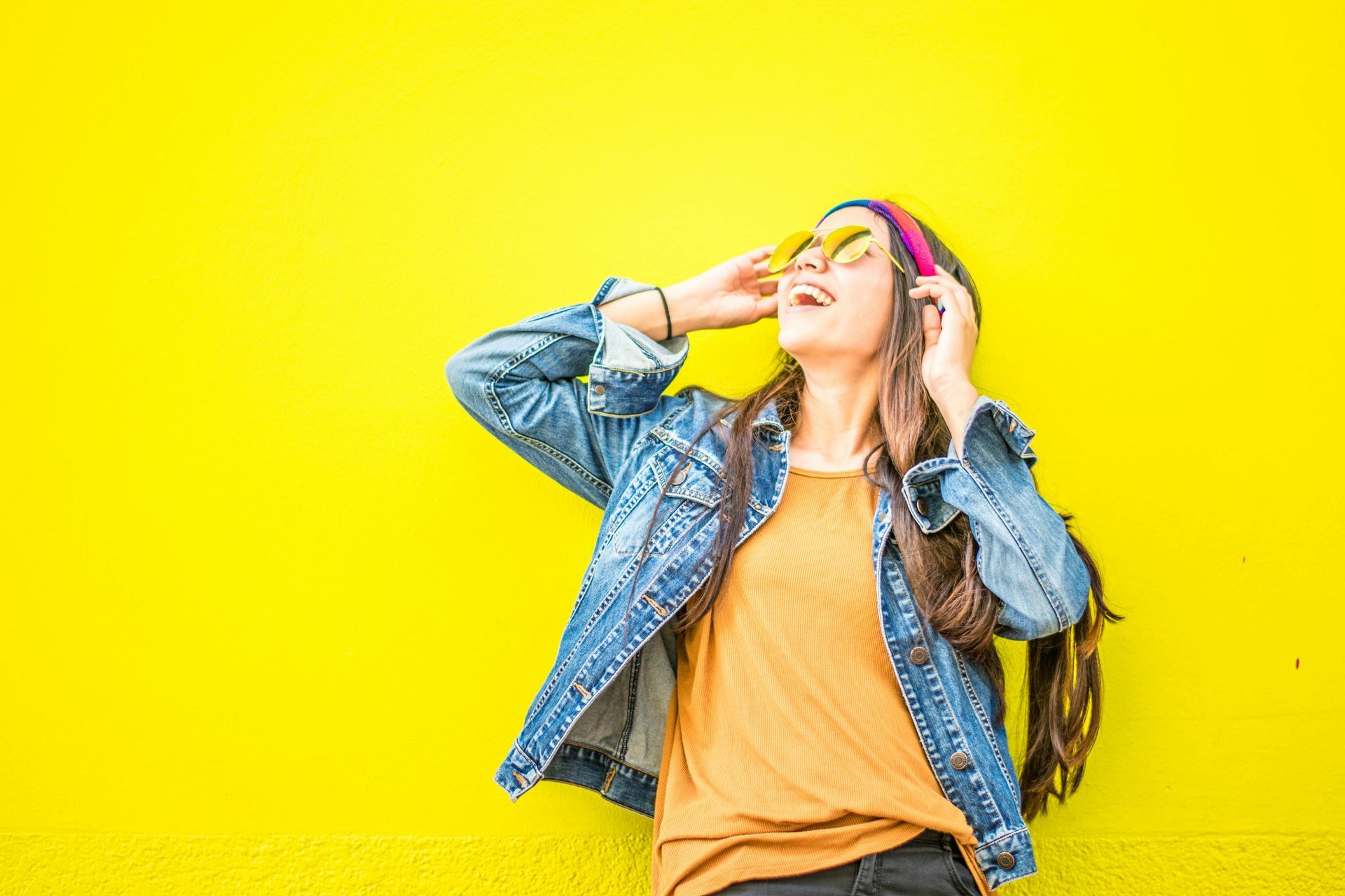 Woman in denim jacket against bright yellow wall, holding sunglasses and looking up