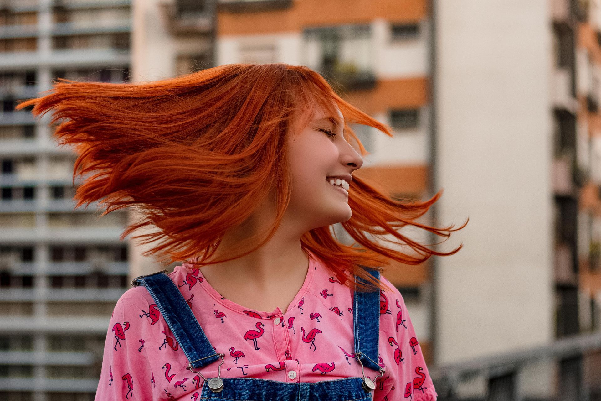A woman with red hair is standing in front of a building with her hair blowing in the wind.