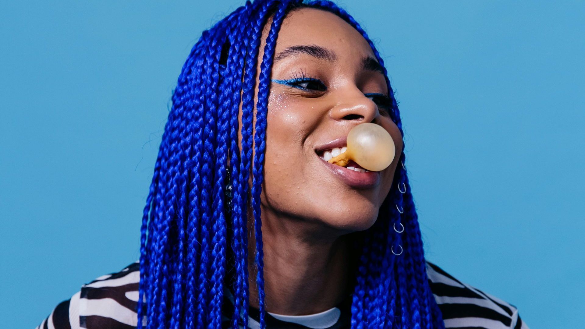 A woman with blue braids is blowing a bubble of gum.