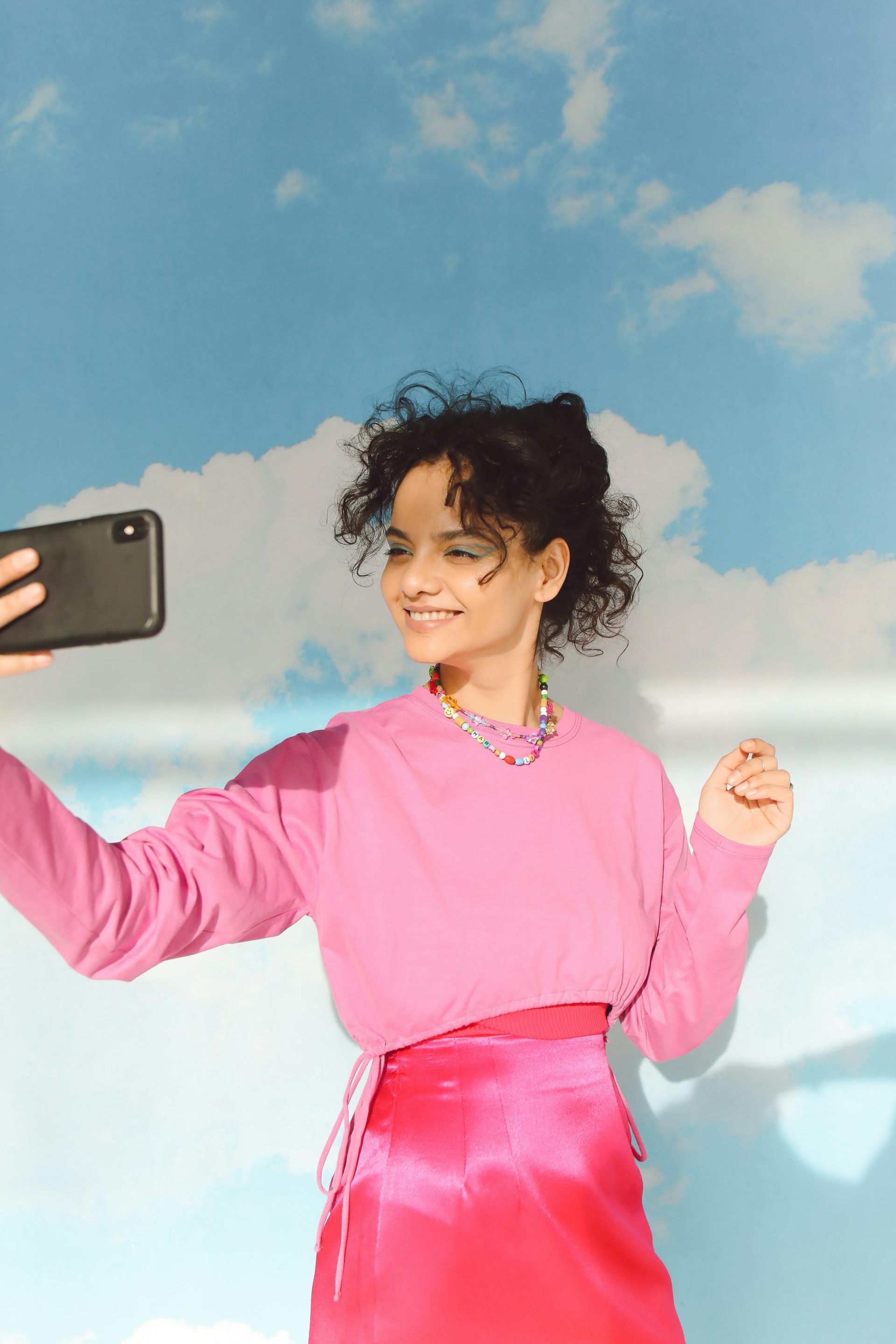 A woman in a pink sweater and pink skirt is taking a selfie with her phone.