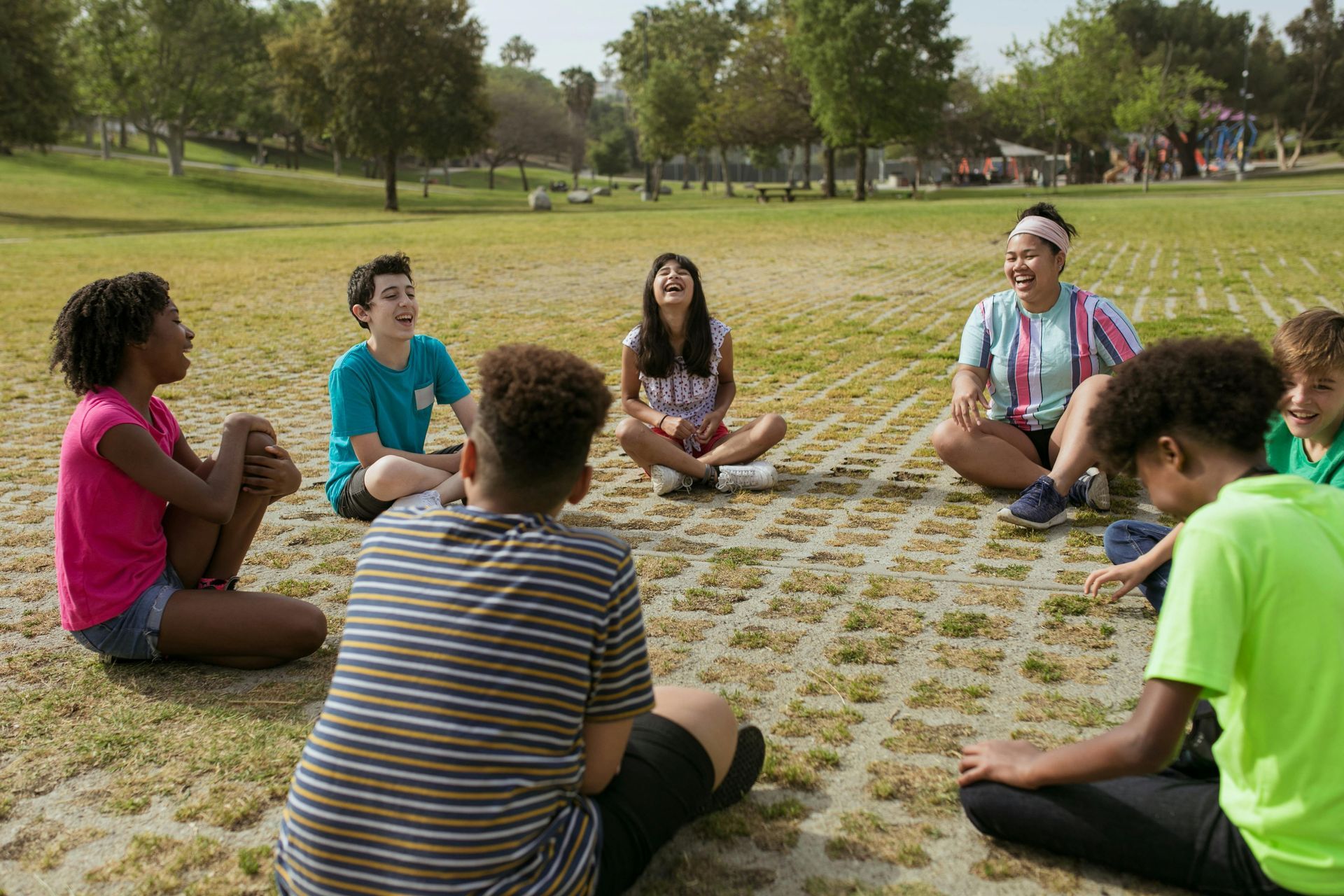 A group of young people are sitting in a circle on the ground in a park.