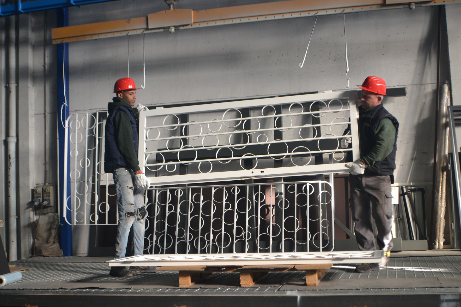 Due uomini che indossano caschi protettivi trasportano una ringhiera metallica su un pallet.