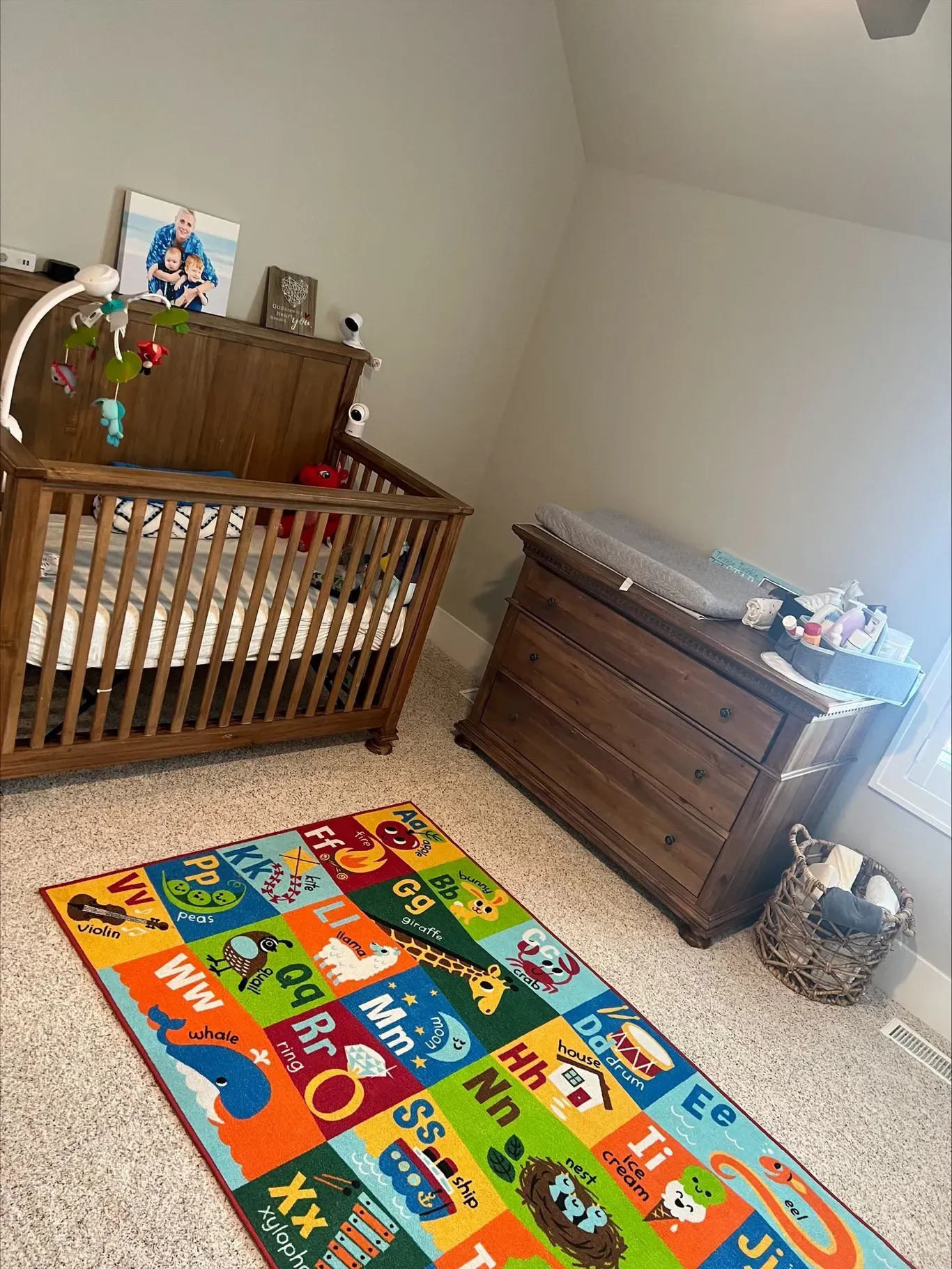 Nursery with wooden crib and dresser, colorful alphabet rug, changing pad, and basket of supplies.
