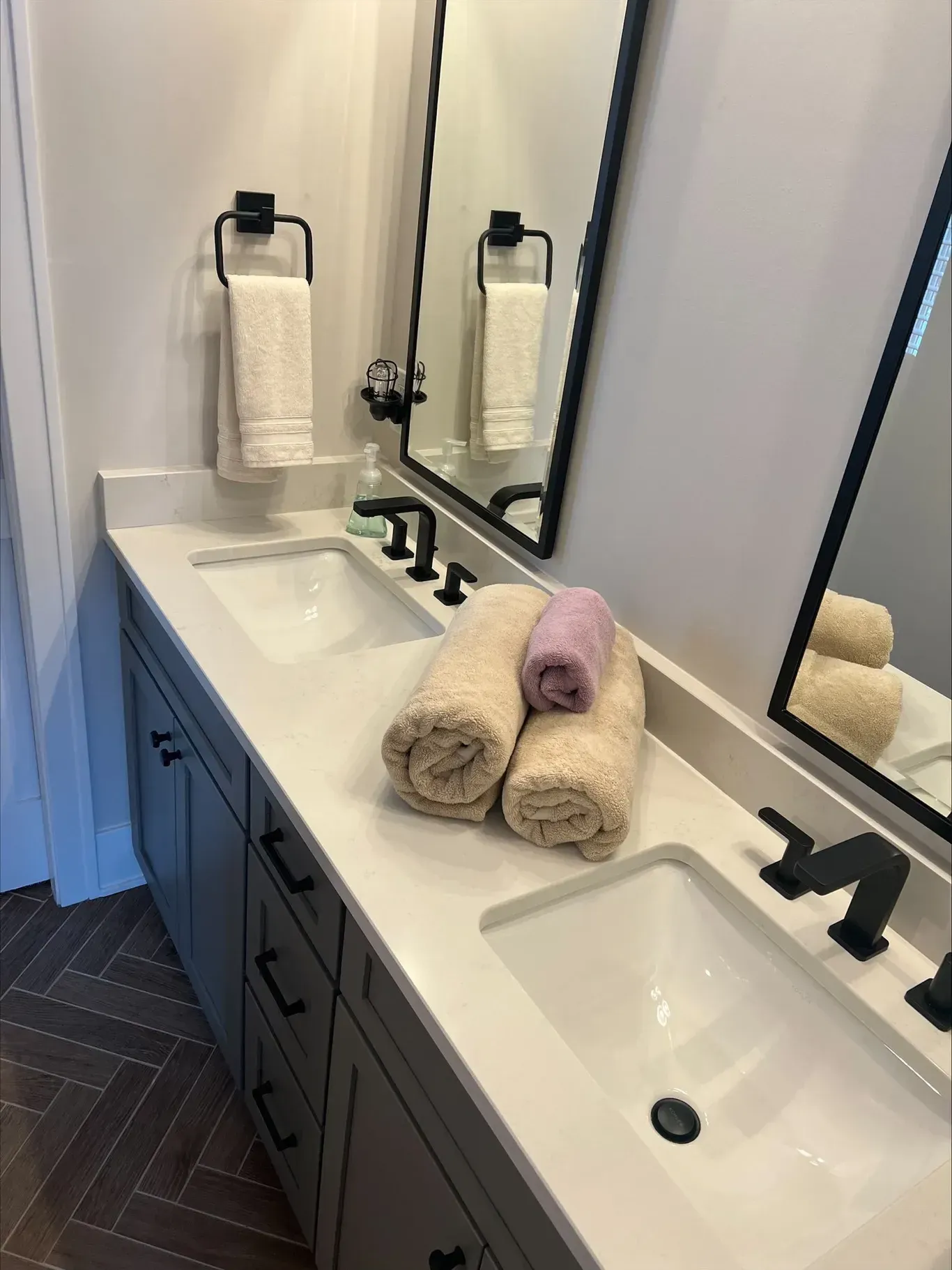 Bathroom with dual sinks, gray cabinets, white countertop, black fixtures, and rolled towels.