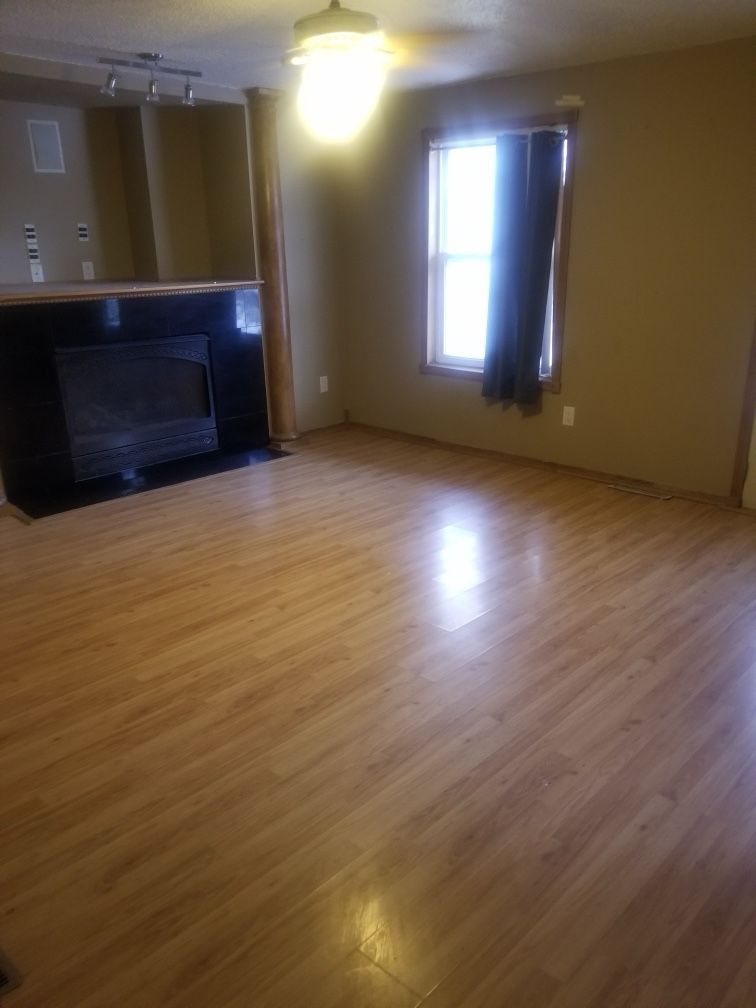 Empty living room with wood-look flooring, fireplace, window with curtain, and a ceiling fan.