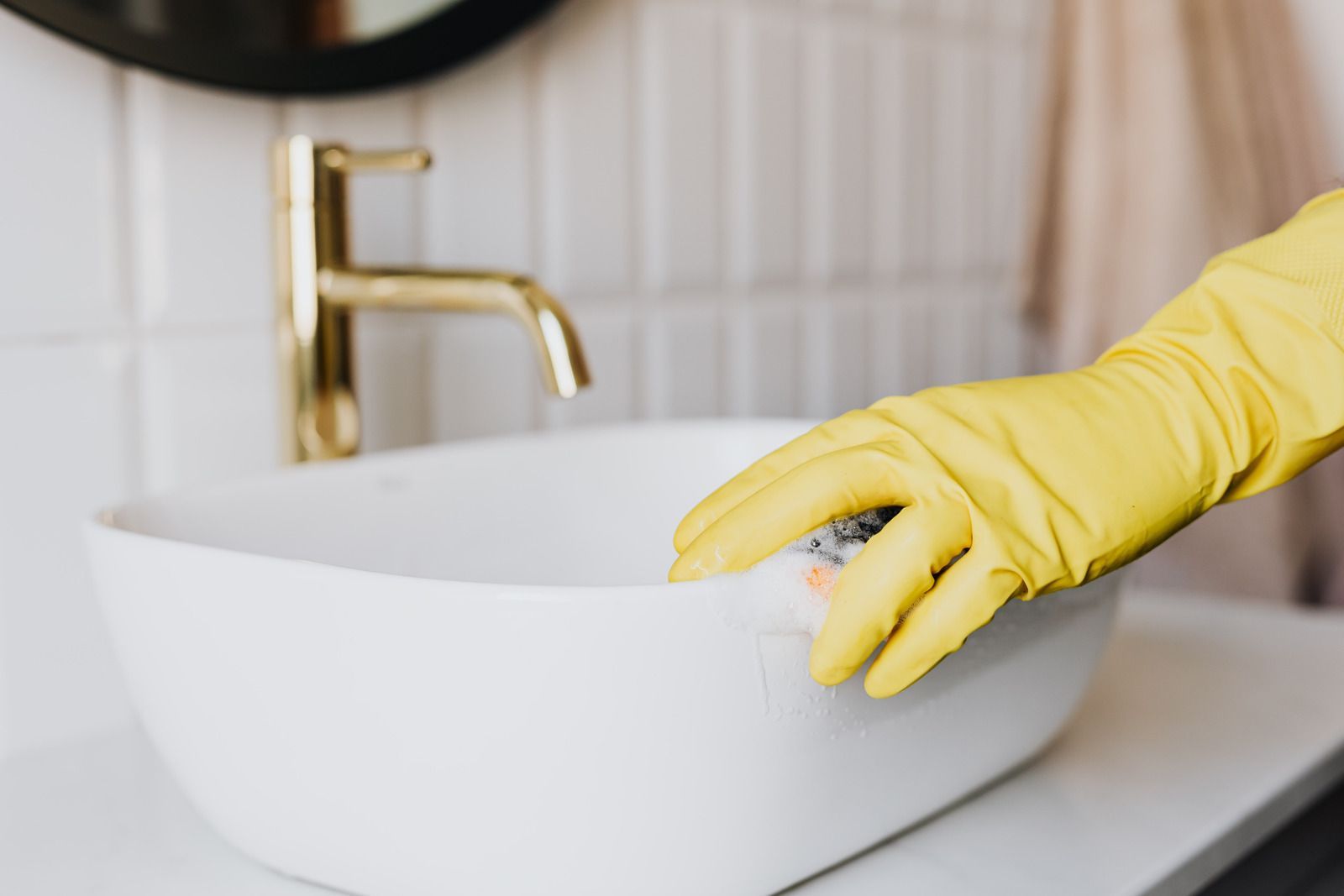 A person wearing yellow rubber gloves is cleaning a bathroom sink with a sponge.
